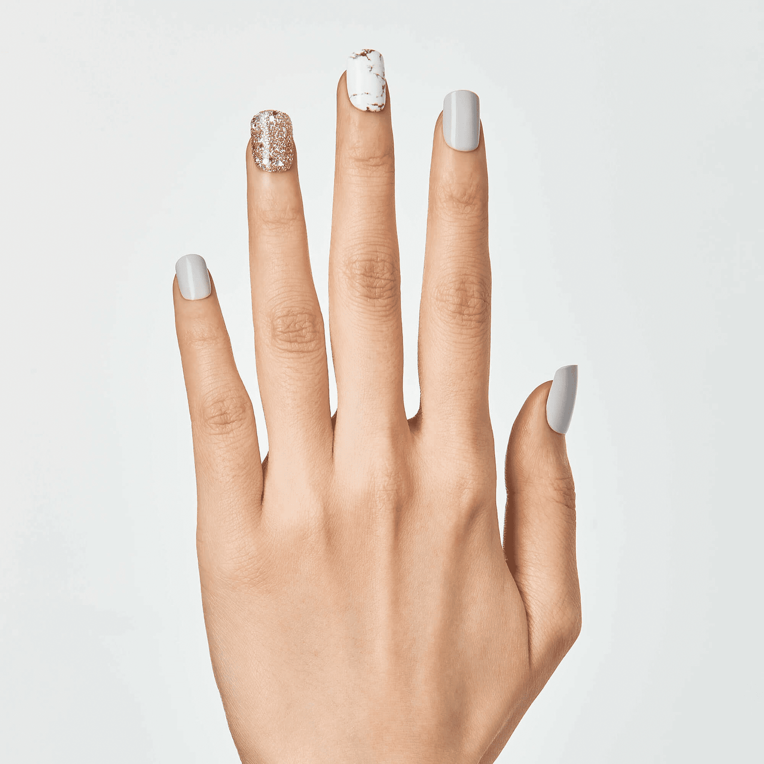 Hand with medium-length press-on nails featuring a gray matte finish, one glittery rose gold nail, and one white nail with gold marble accents.