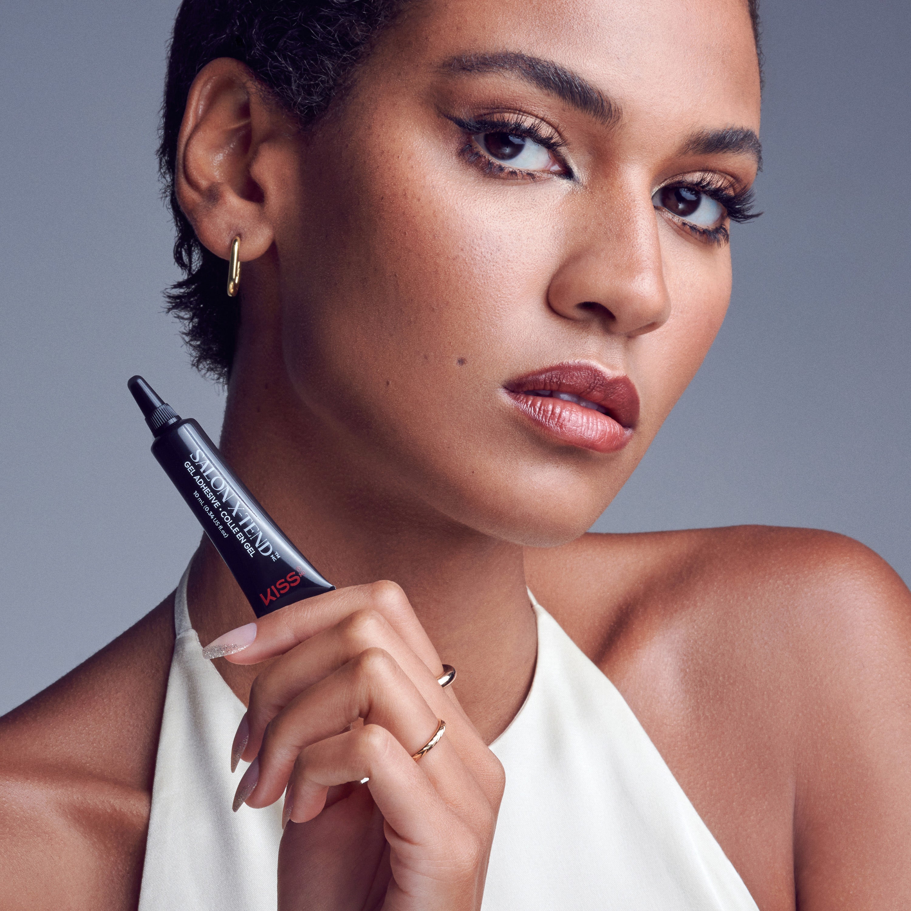 Close-up of a woman with natural makeup and gold hoop earrings holding a black tube of KISS Salon X-tend Gel Adhesive with white text and a red logo, against a gray background.
