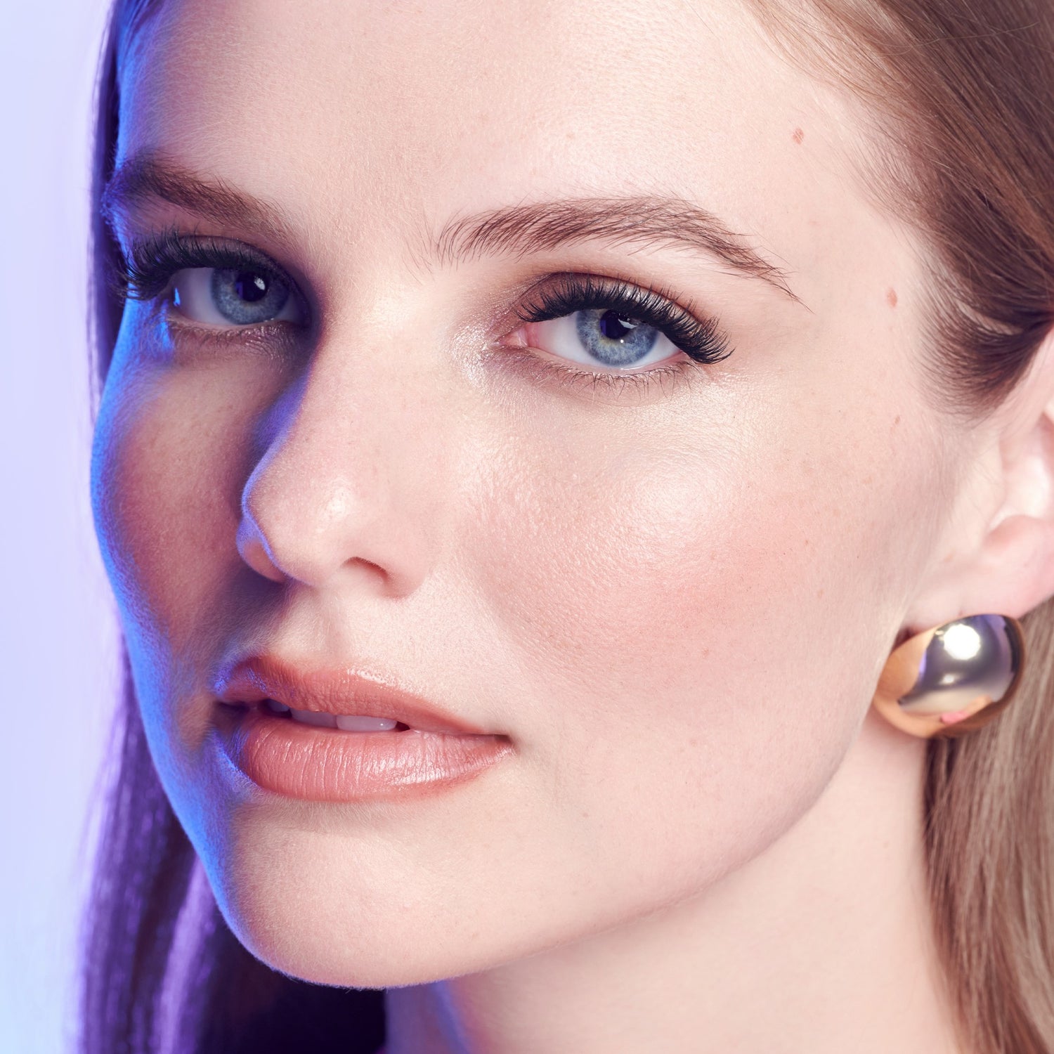 Woman with blue eyes and brown hair wearing gold hoop earrings, purple-tinted lighting.