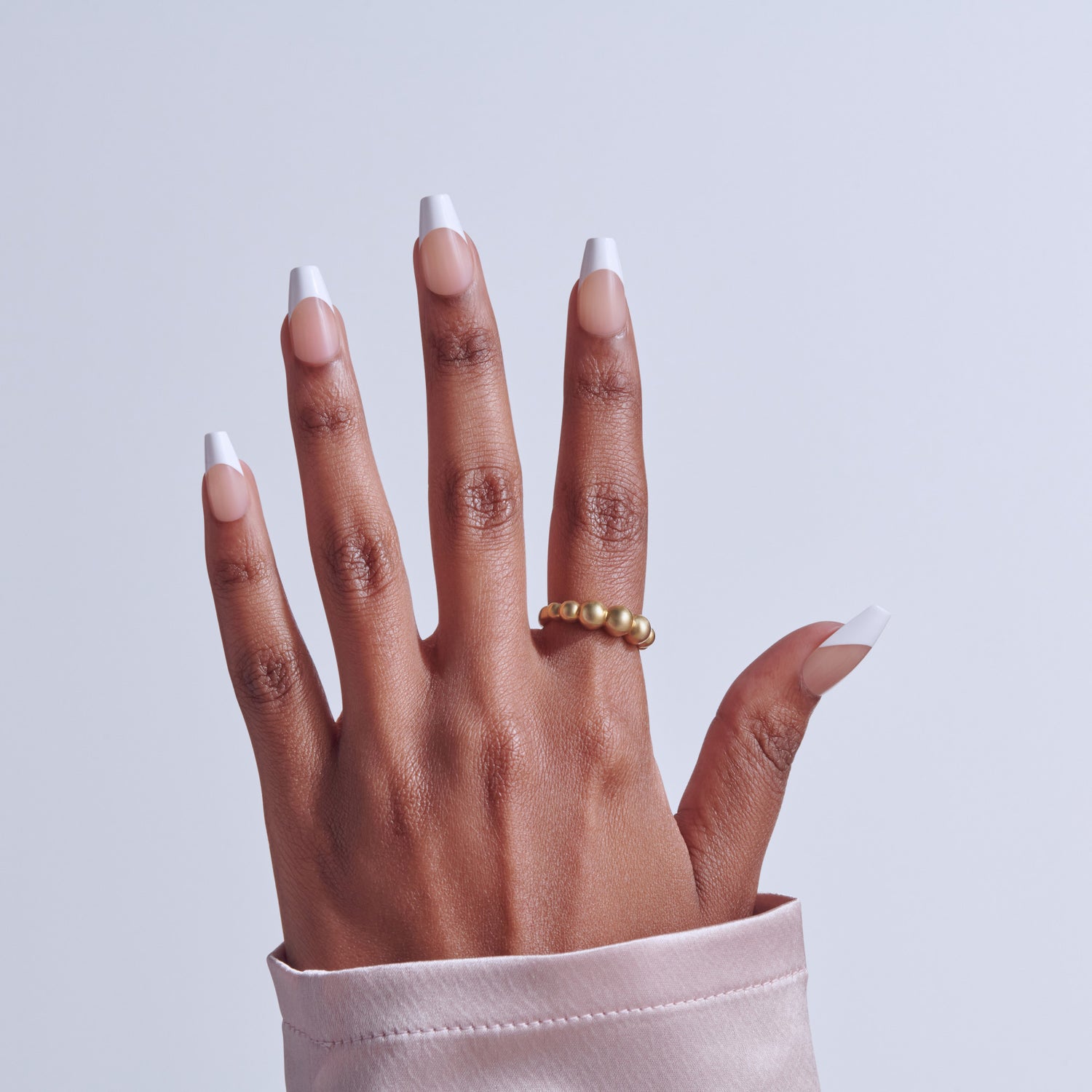 Hand with white-tipped acrylic nails and gold beaded ring against light background.