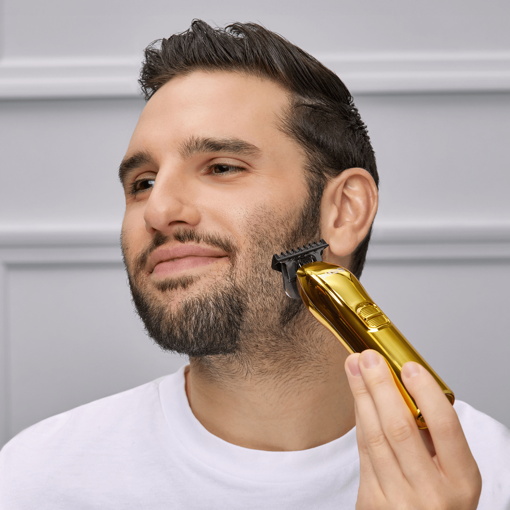 Man with dark hair and beard using a shiny gold cordless T-blade trimmer on his cheek against a light gray background.