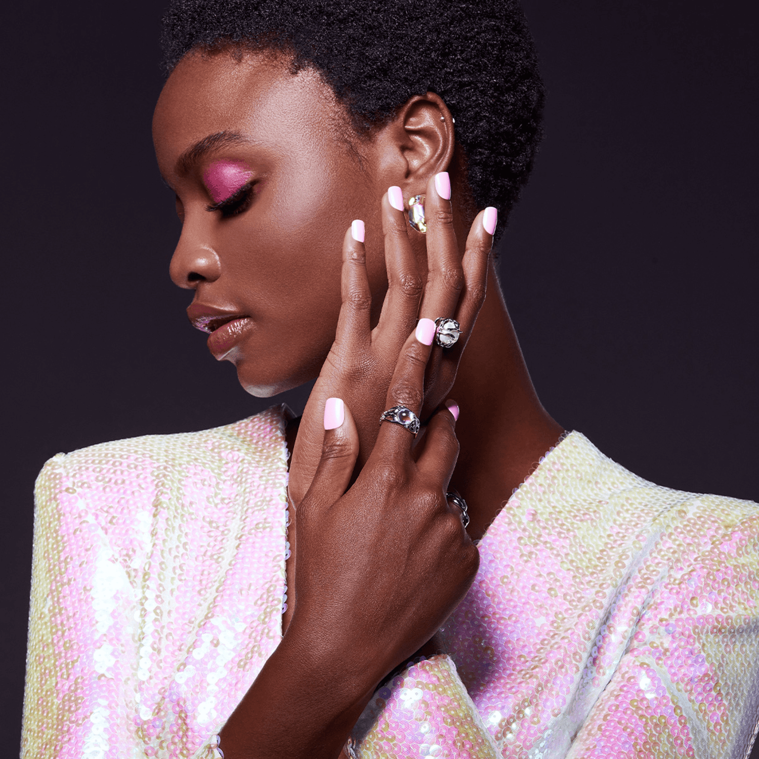 Woman with short curly hair wearing shiny pink and white sequin jacket, showcasing glossy pastel pink press-on nails and silver rings on dark background.