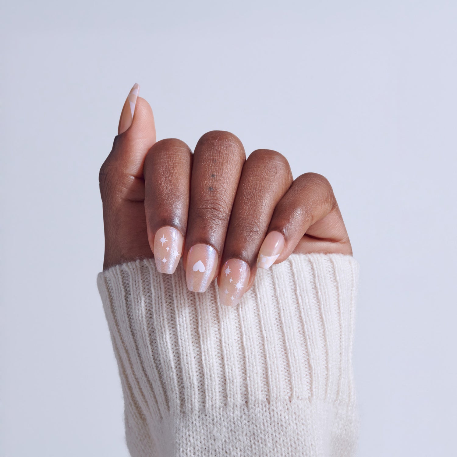 Hand with nude nail polish featuring white stars and heart designs, wearing cream sweater.
