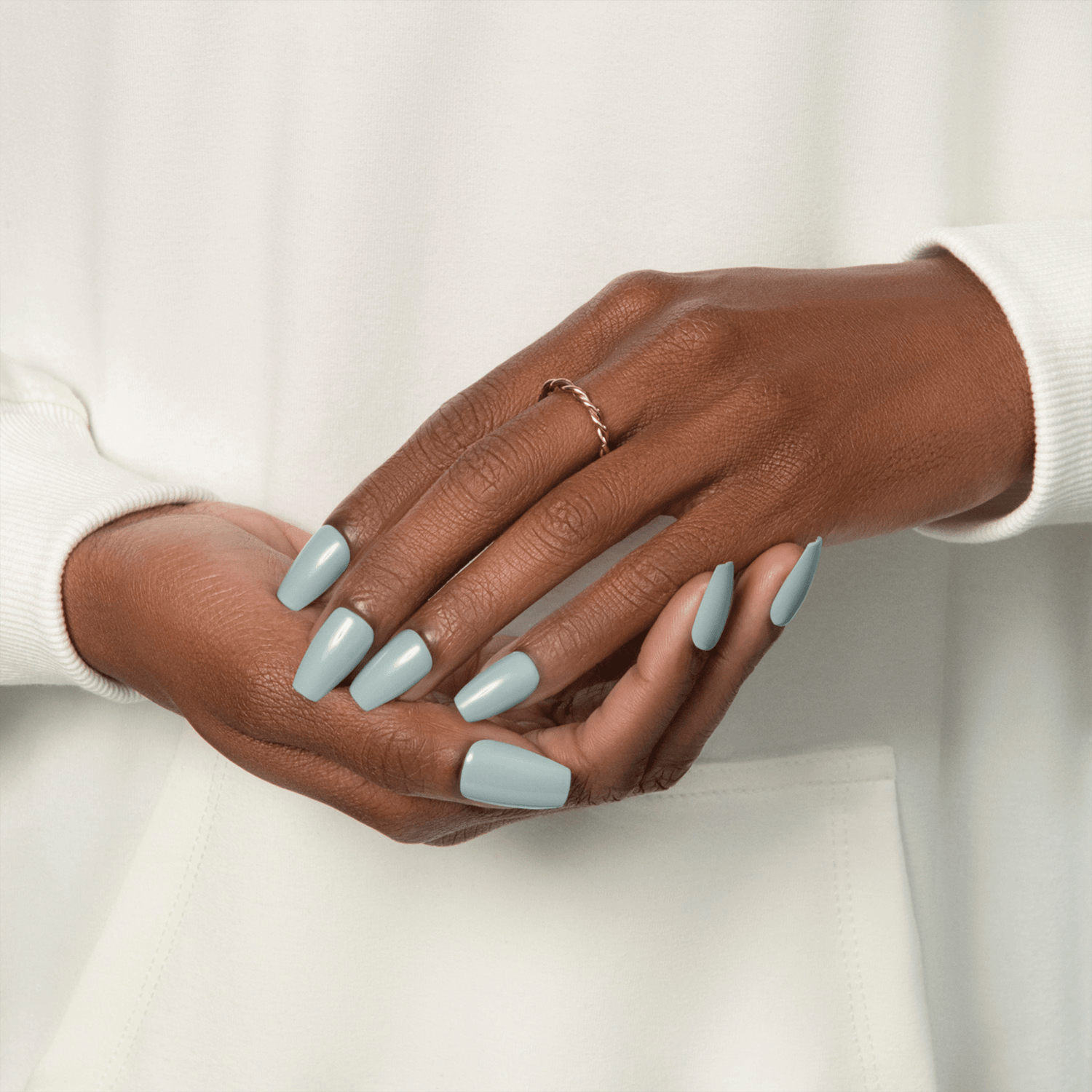 Hands with medium brown skin tone wearing long, glossy muted green press-on nails, styled with a simple twisted ring and white long-sleeve top.