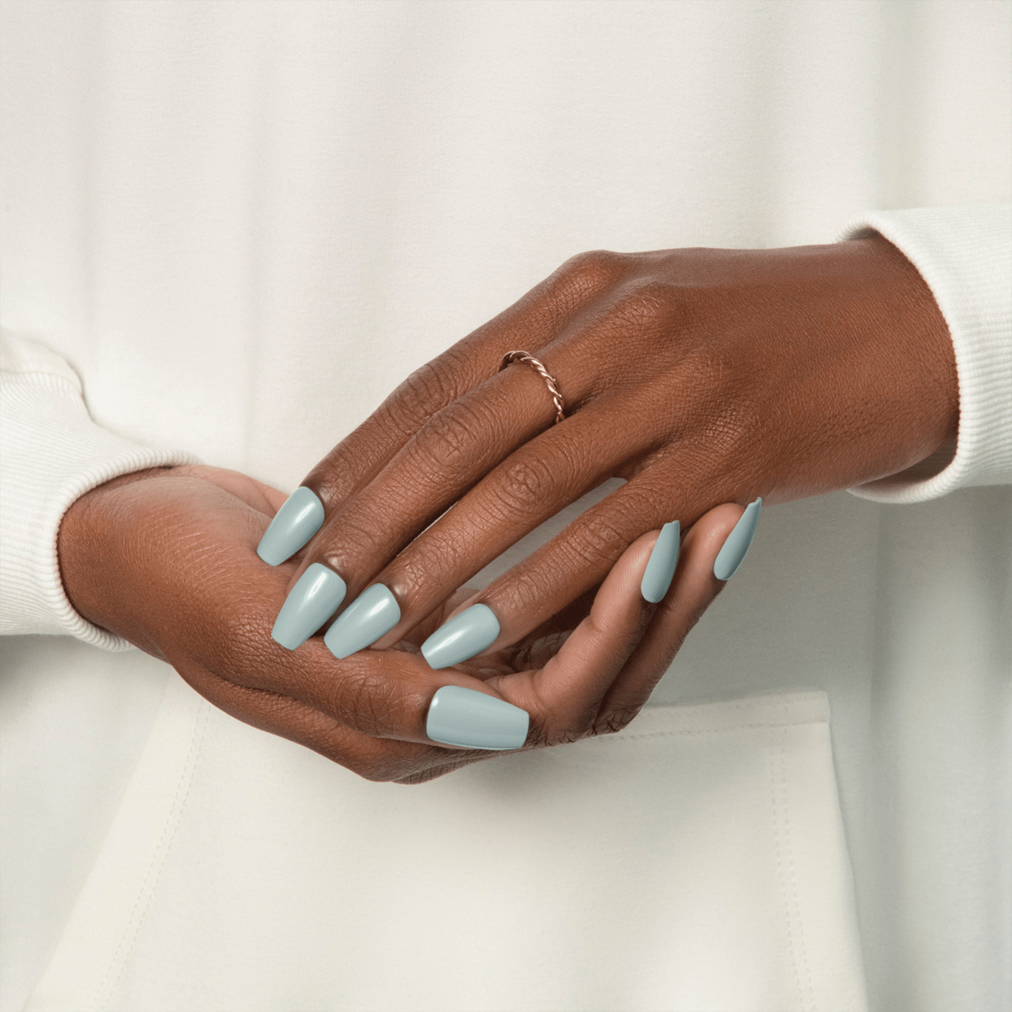 Hands with medium brown skin tone wearing long, glossy muted green press-on nails, styled with a simple twisted ring and white long-sleeve top.