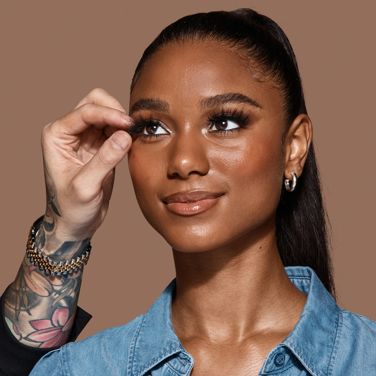 Close-up of a smiling woman with dark skin and sleek pulled-back hair having long, voluminous strip lashes applied by a tattooed hand, wearing a blue denim shirt.