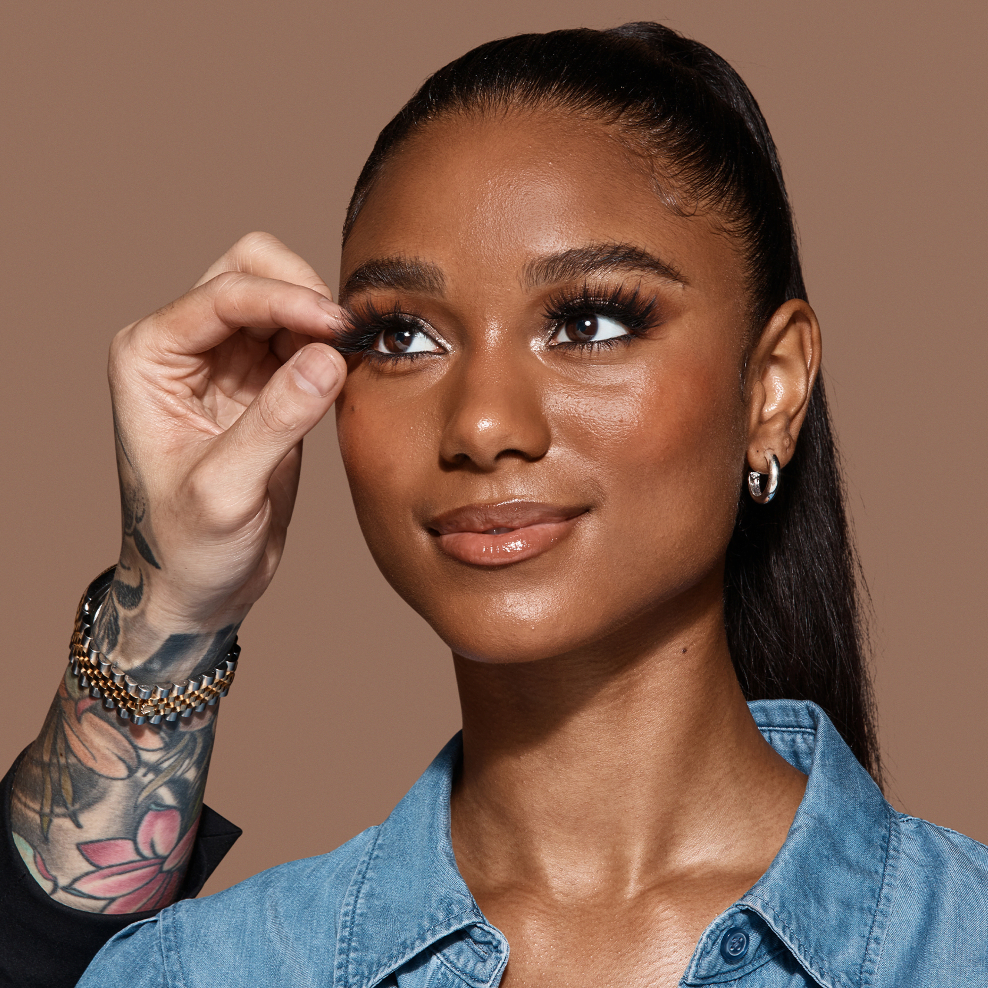 Close-up of a smiling woman with dark skin and sleek pulled-back hair having long, voluminous strip lashes applied by a tattooed hand, wearing a blue denim shirt.