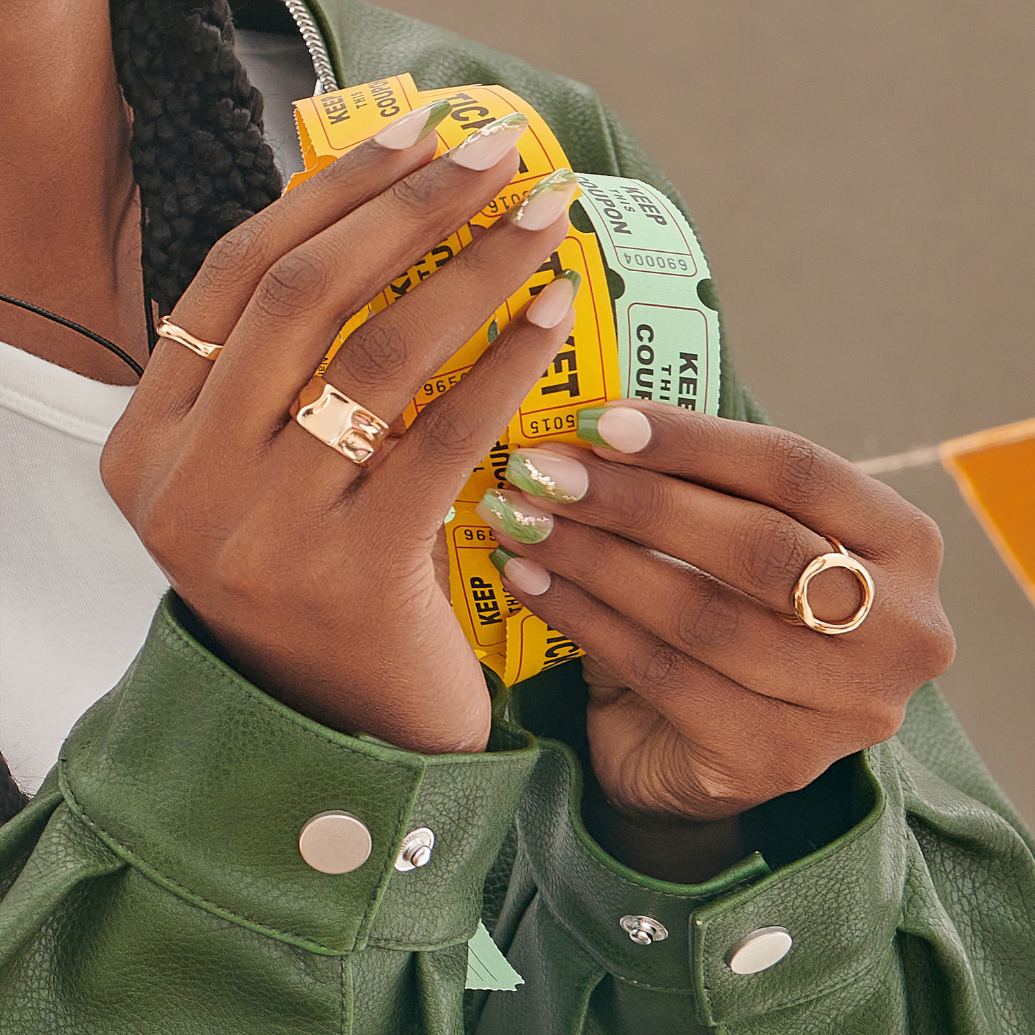 Hands with matte ballerina-shaped press-on nails featuring a soft nude base and sparkly green tips, holding yellow and green raffle tickets.