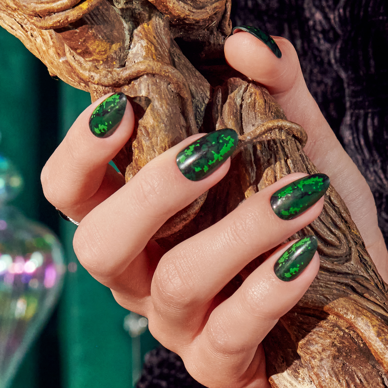 Hand with almond-shaped nails painted dark green with bright green speckles, holding a carved wooden object against a dark textured background.