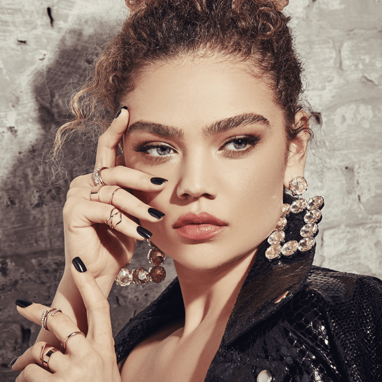 Woman with curly hair wearing large crystal earrings and a black textured jacket, showing glossy black press-on nails on her fingers adorned with several gold rings.