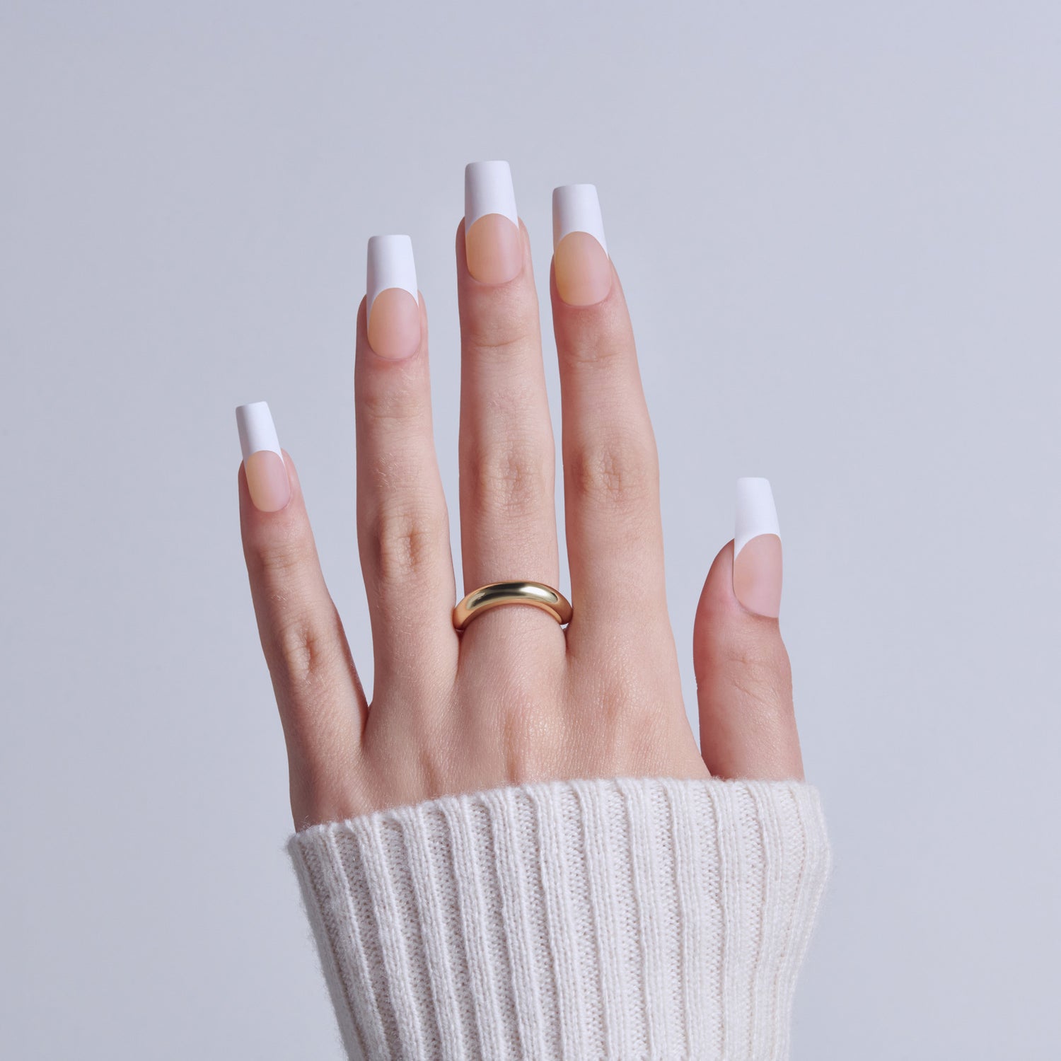 Hand with classic French manicure nails and gold band ring, white sweater cuff visible.