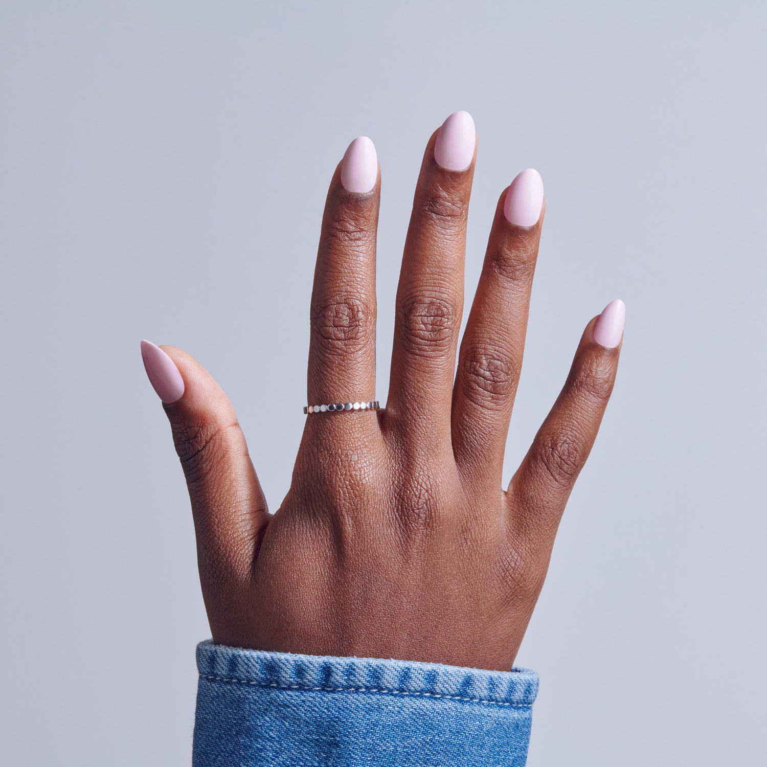 Hand with pale pink oval nails and silver ring against blue cuff on gray background.