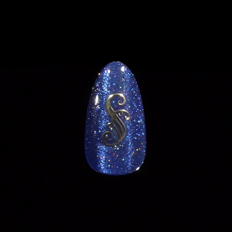 Single artificial nail tip painted with glittery deep blue polish, featuring a small metallic gold ornate \