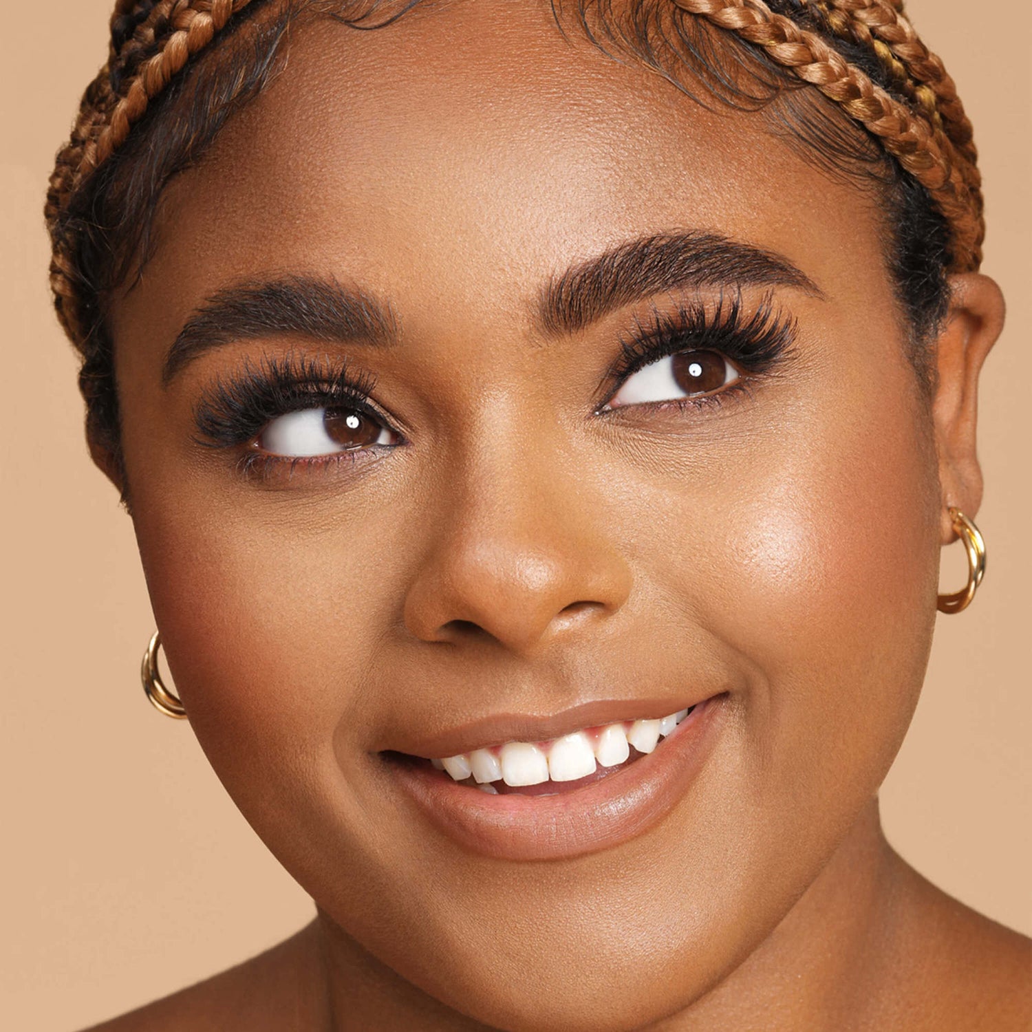 Close-up of a smiling woman with braided hair wearing thick, voluminous false eyelashes and gold hoop earrings against a beige background.