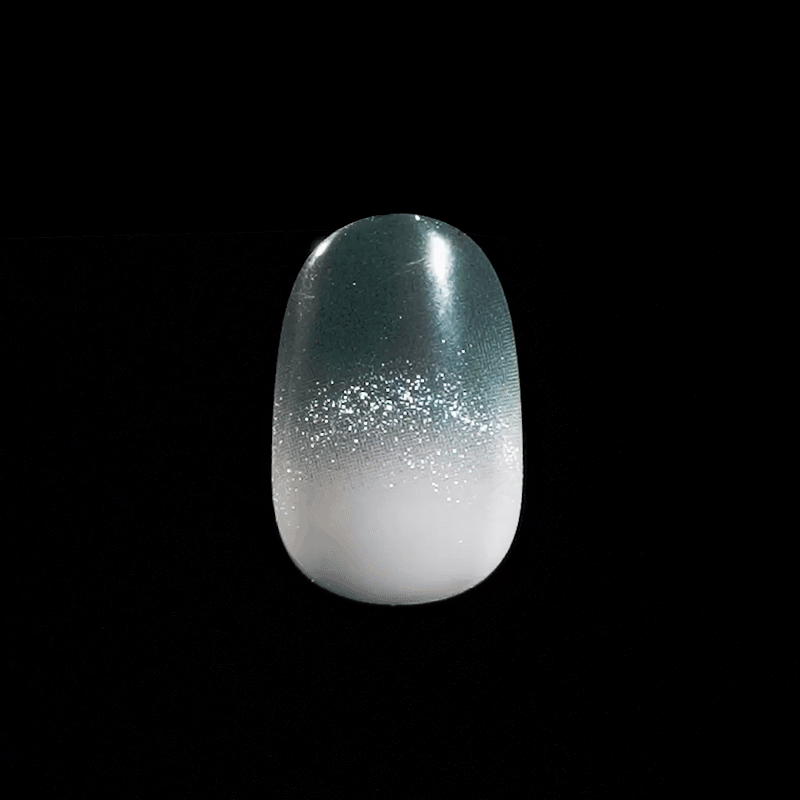 Single artificial nail in close-up with an ombré design transitioning from dark teal at the top to white at the base, featuring fine silver glitter in the middle.