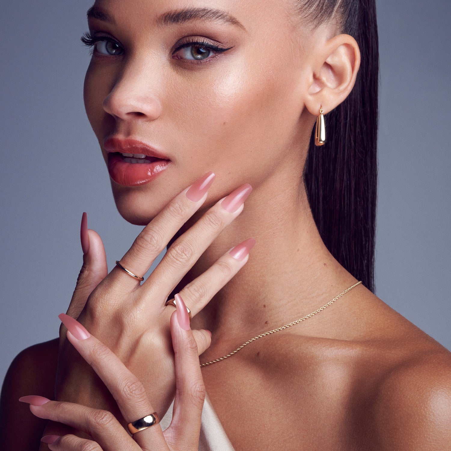 Woman with smooth skin and glossy pink eyeshadow, lips, and long square-shaped soft gel nails in a muted rose tone, wearing gold jewelry on hands and ears.
