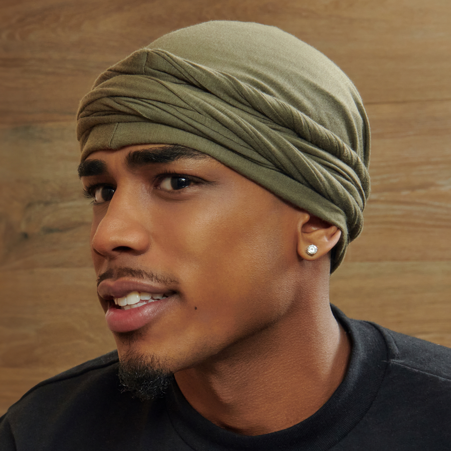 Close-up side view of a young man wearing an olive green satin-lined halo turban with twisted fabric detail, set against a wooden background.