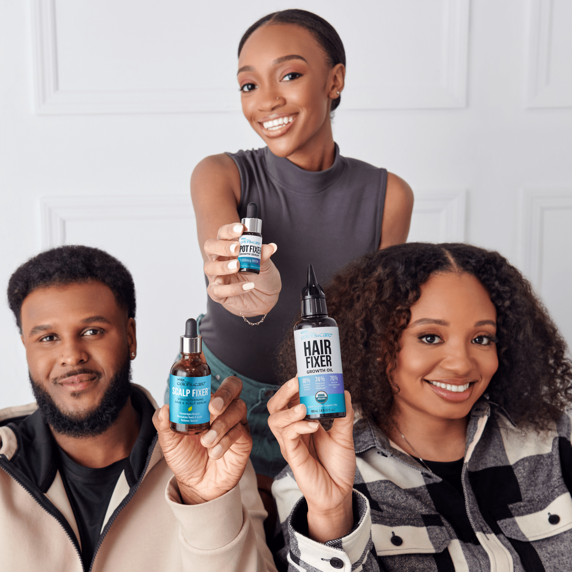 Three smiling people hold KISS Colors &amp; Care hair growth products in blue-labeled bottles: Scalp Fixer serum, Spot Fixer growth serum, and Hair Fixer growth oil.