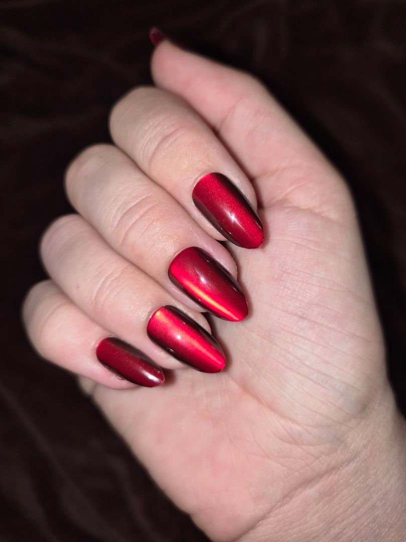 Red metallic almond-shaped nails with chrome finish on pale hand against dark background.