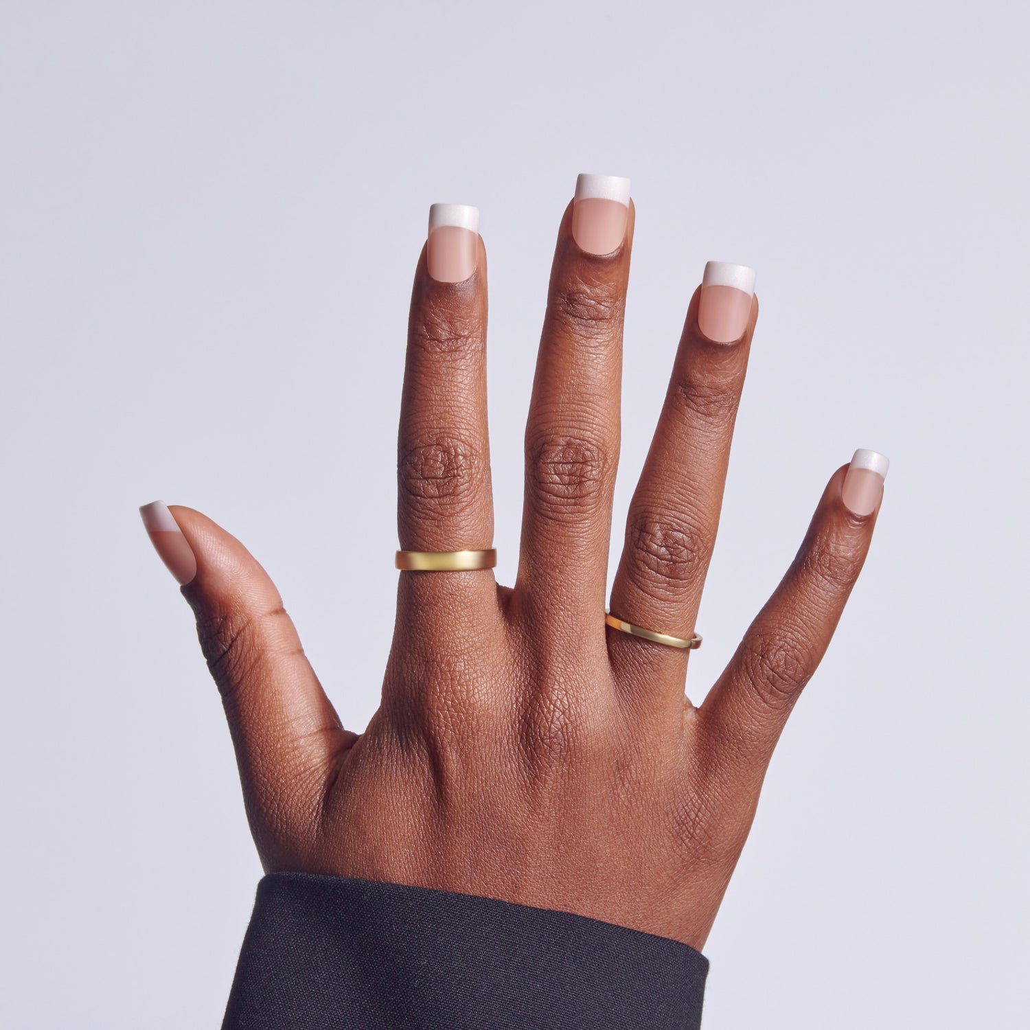 Hand with white-tipped manicure and gold rings against light background.