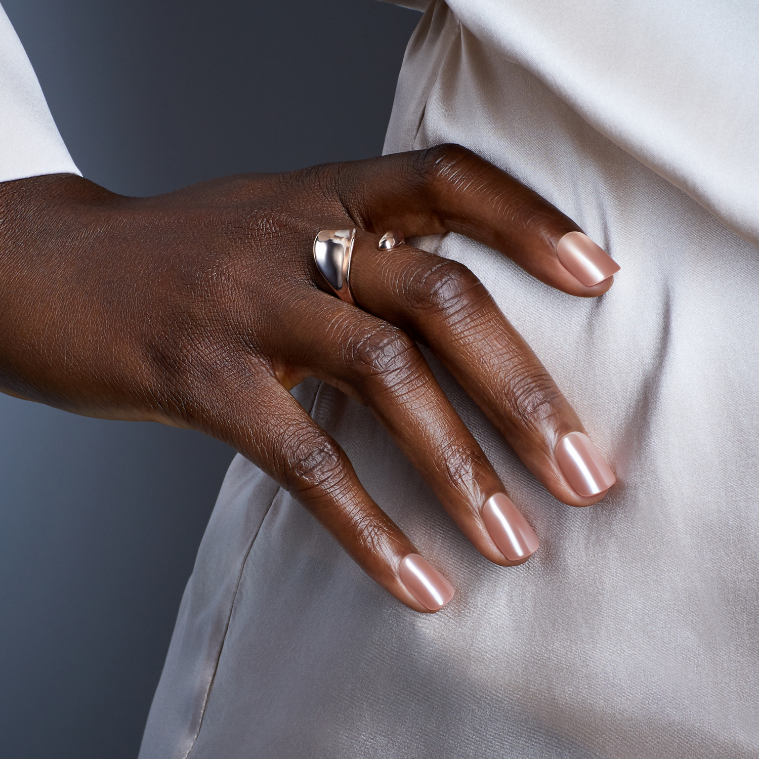 Hand with medium brown skin tone wearing metallic rose gold press-on nails, gently touching the side of a light champagne satin fabric, with a smooth, glossy finish.