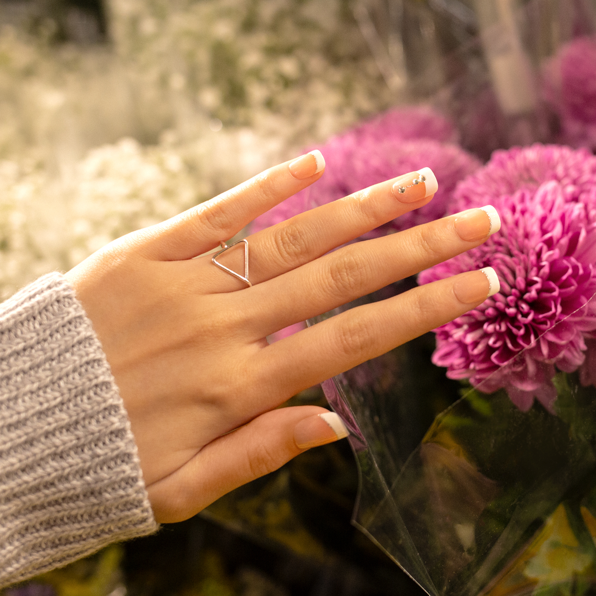 Hand with imPRESS press-on nails featuring nude base and white French tips, one nail decorated with silver rhinestones, against a background of purple flowers.
