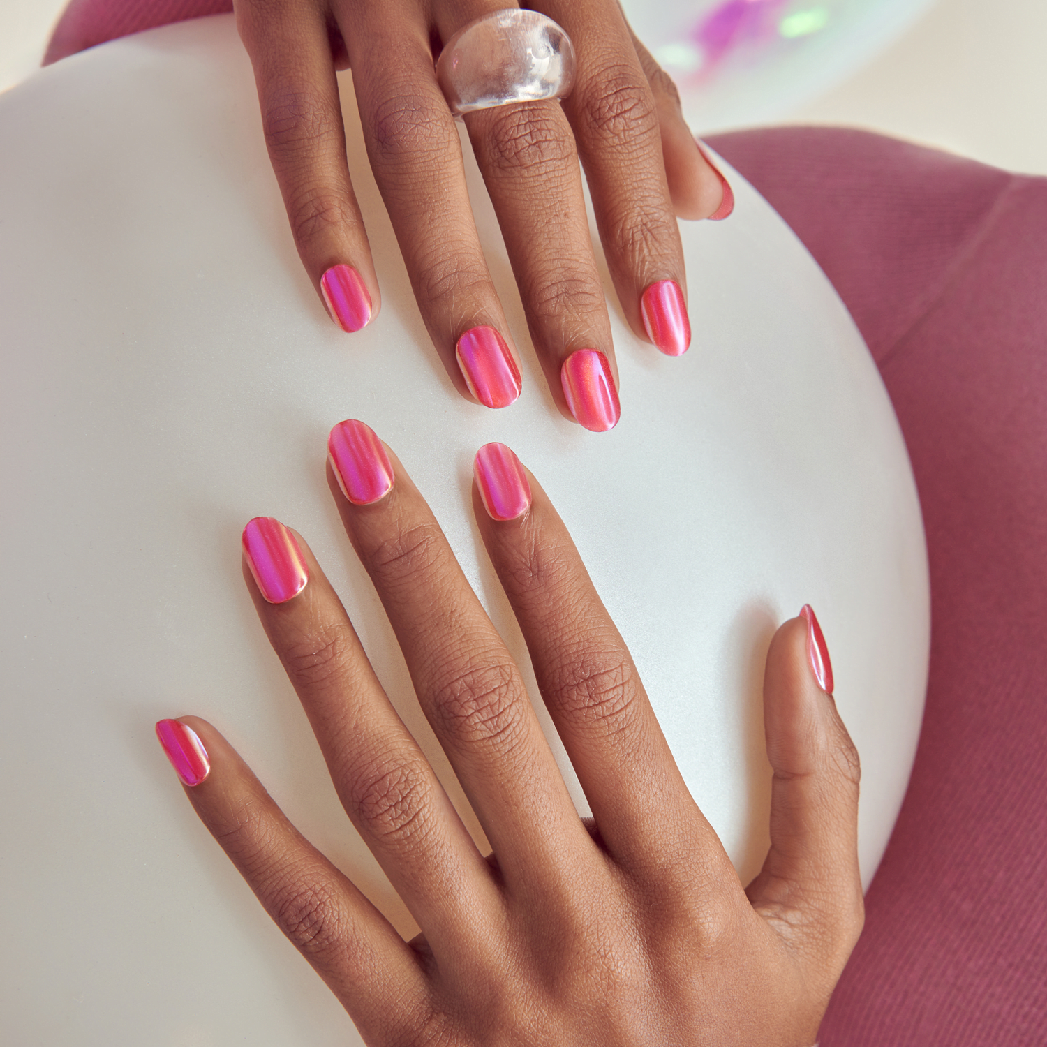 Two hands with medium skin tone resting on a white circular surface, showcasing glossy pink iridescent press-on nails with a rounded shape. One hand wears a clear chunky ring on the middle finger. The background includes a soft purple fabric.