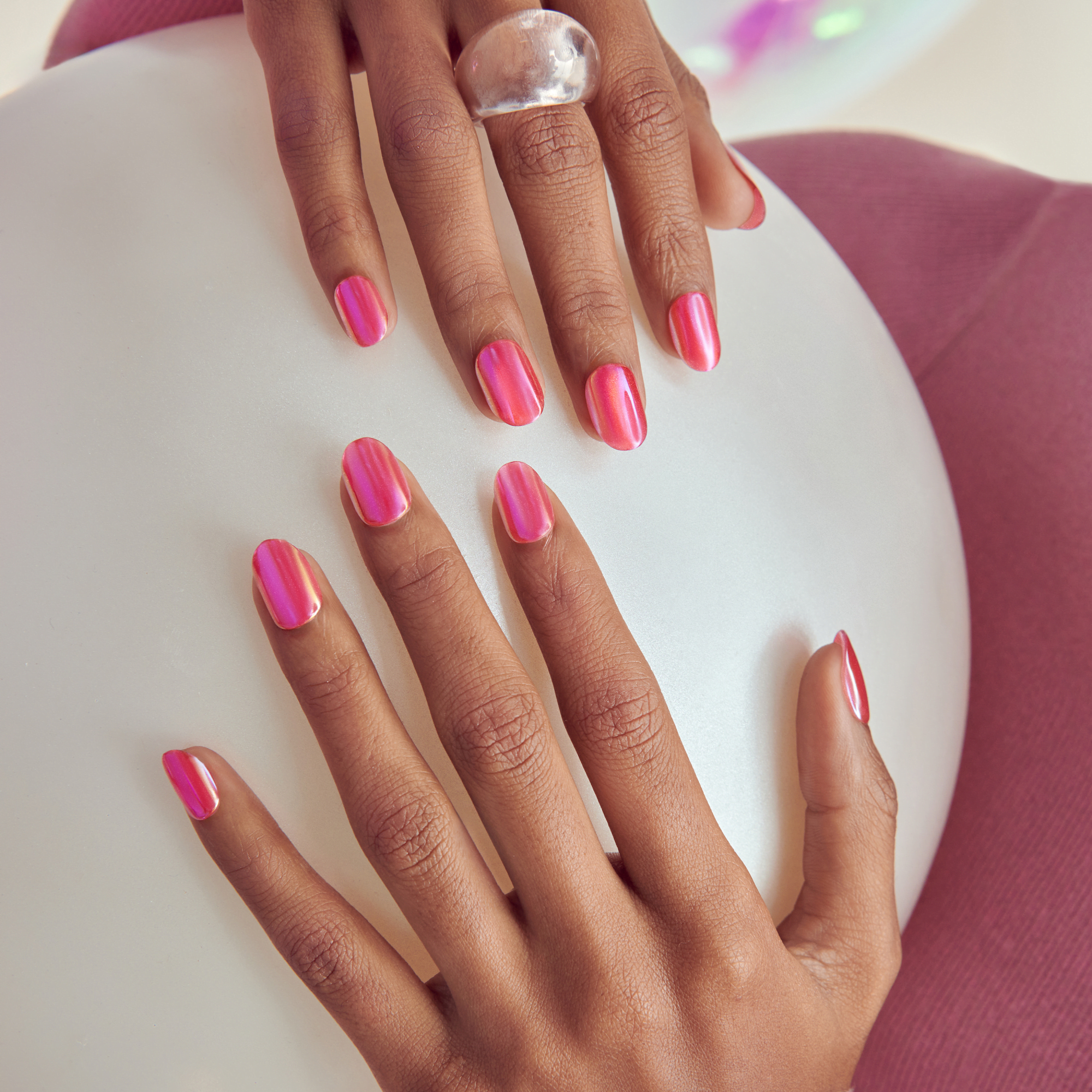 Two hands with medium skin tone resting on a white circular surface, showcasing glossy pink iridescent press-on nails with a rounded shape. One hand wears a clear chunky ring on the middle finger. The background includes a soft purple fabric.