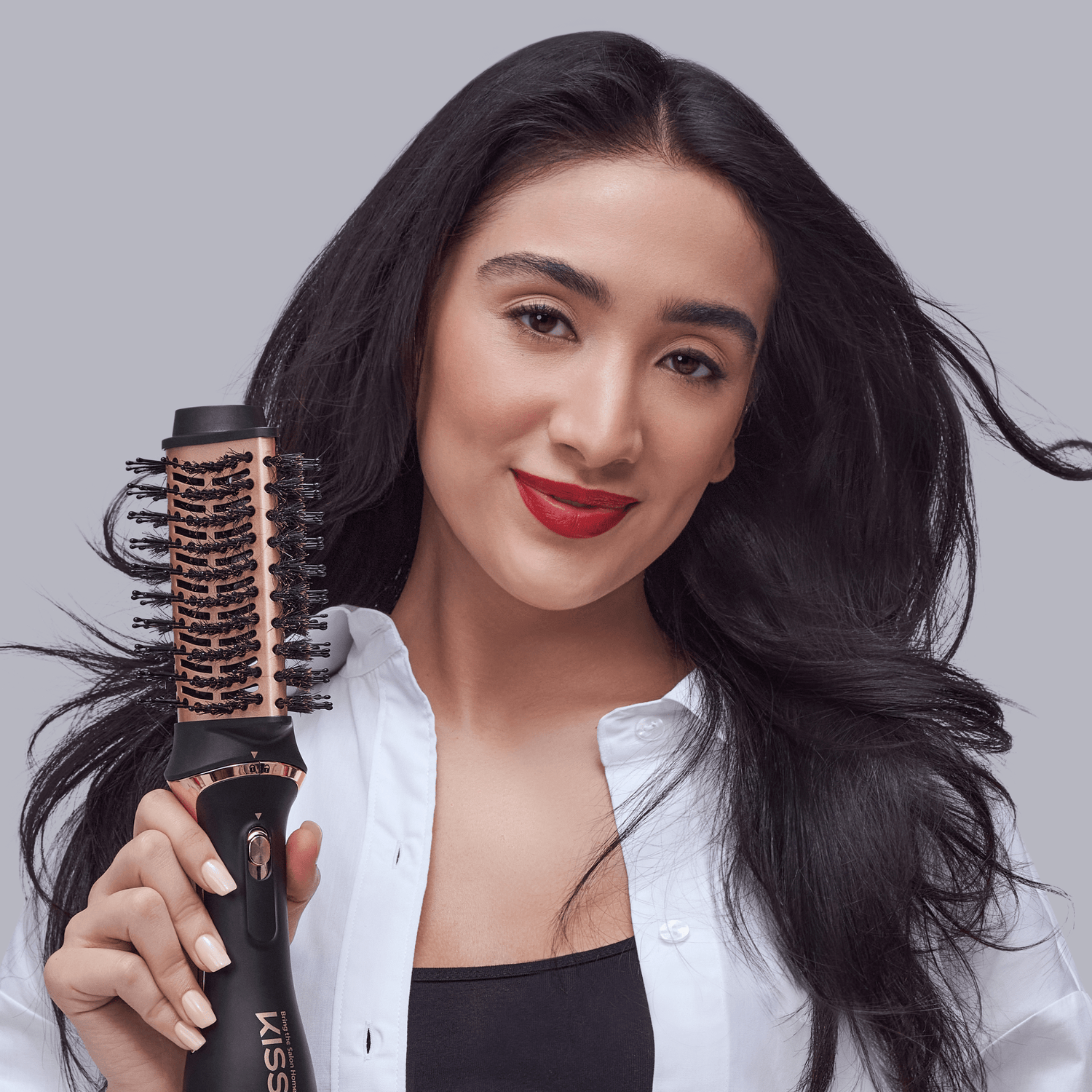 Woman with long black hair and red lipstick holding a black and rose gold KISS Volume &amp; Style Boosting 2-in-1 Air Brush against a light gray background.