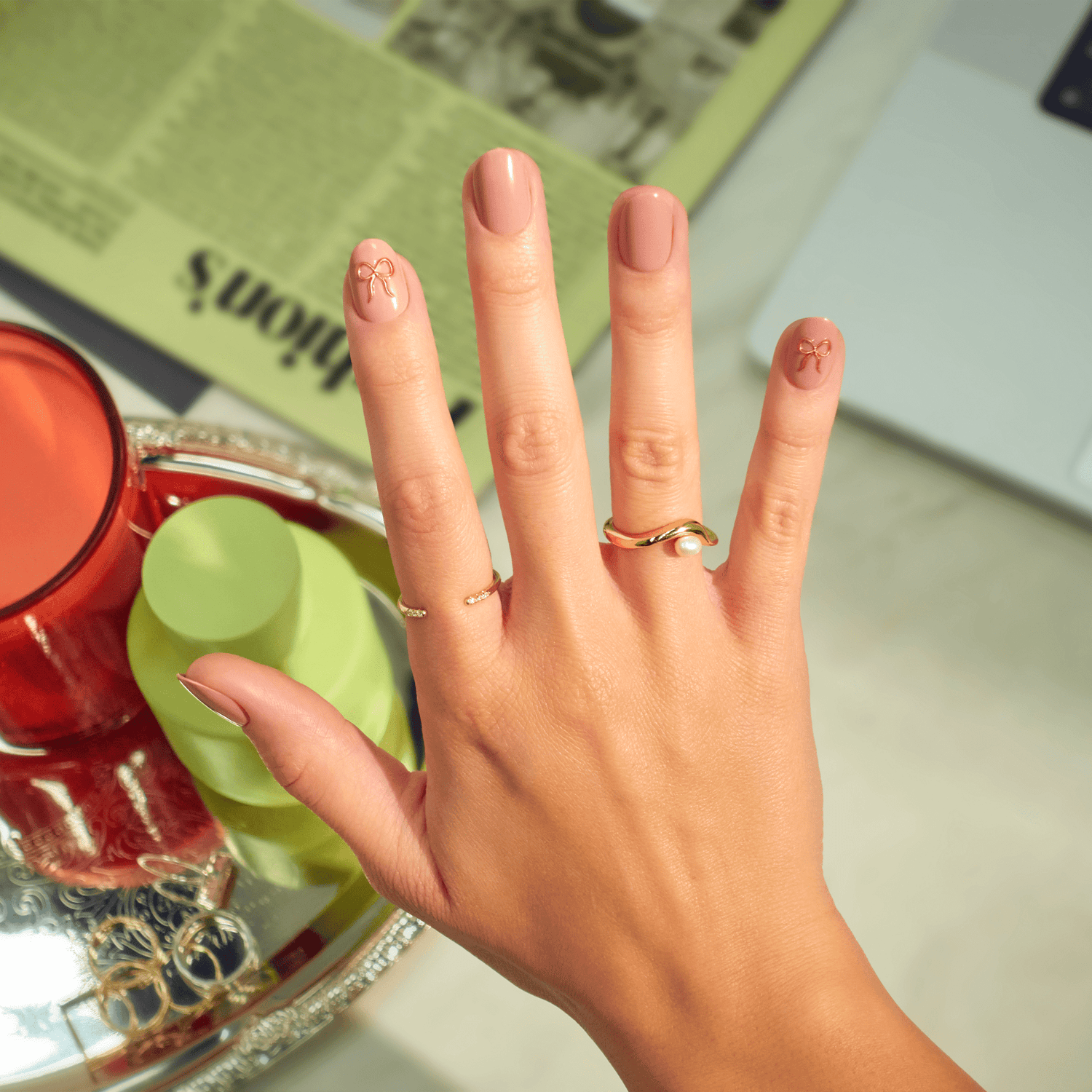 Manicured hand with pink nails featuring bow designs, gold rings, styled flatlay.