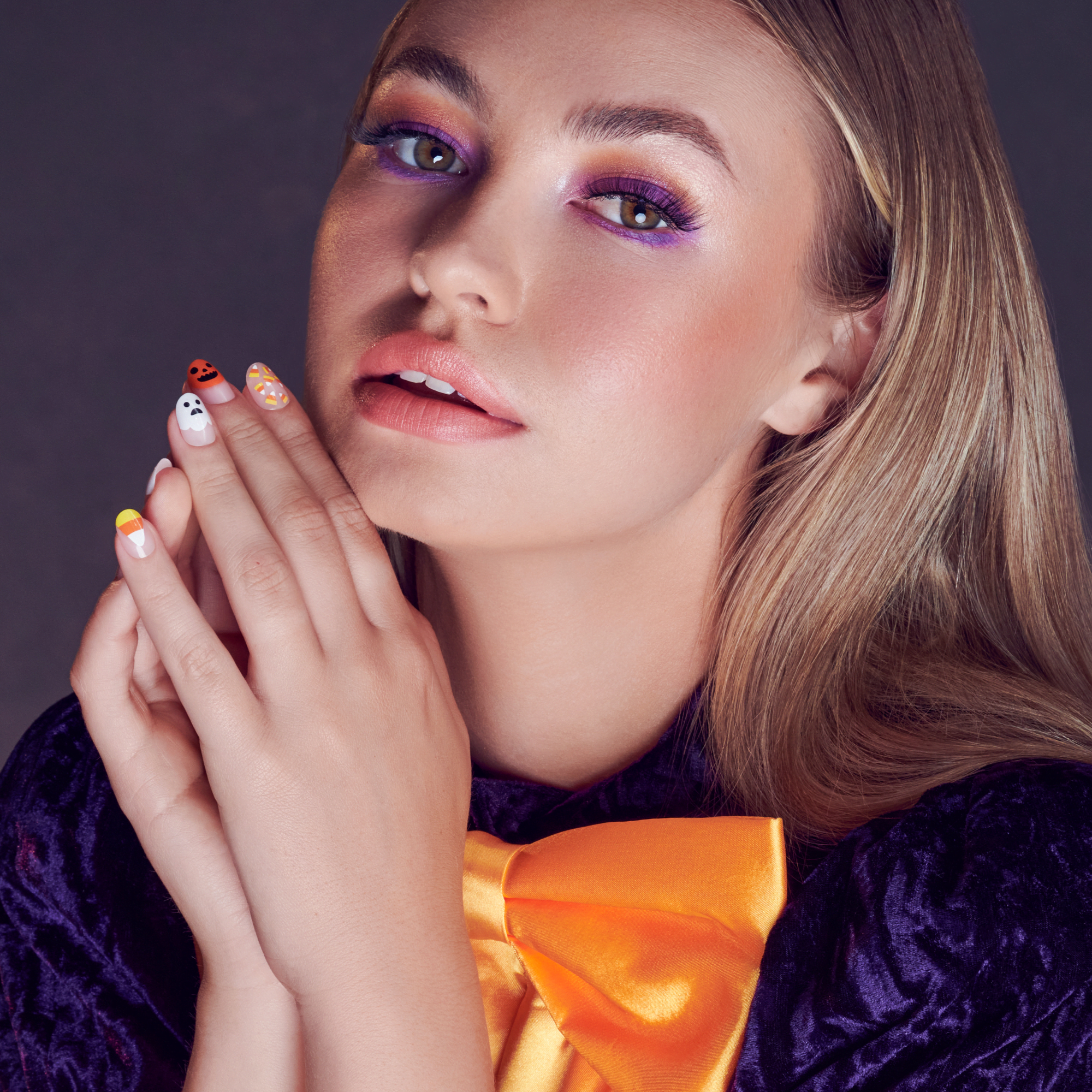 Young woman with light brown hair wearing purple eyeshadow and a large orange satin bow, showing Halloween-themed press-on nails featuring designs like a ghost, jack-o&