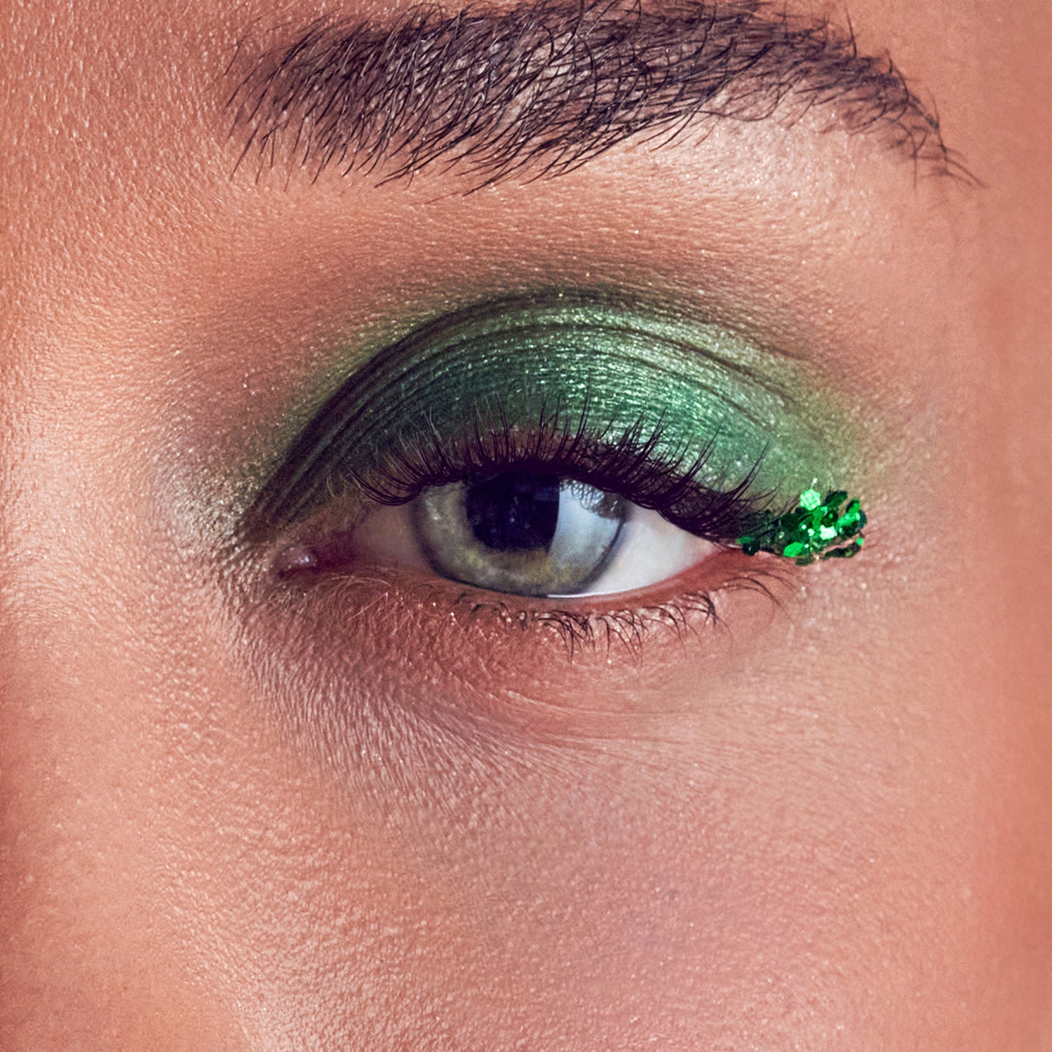 Close-up of an eye with long, dark false eyelashes and shimmering green eyeshadow extending from the eyelid to the outer corner, which is decorated with small green sequins.