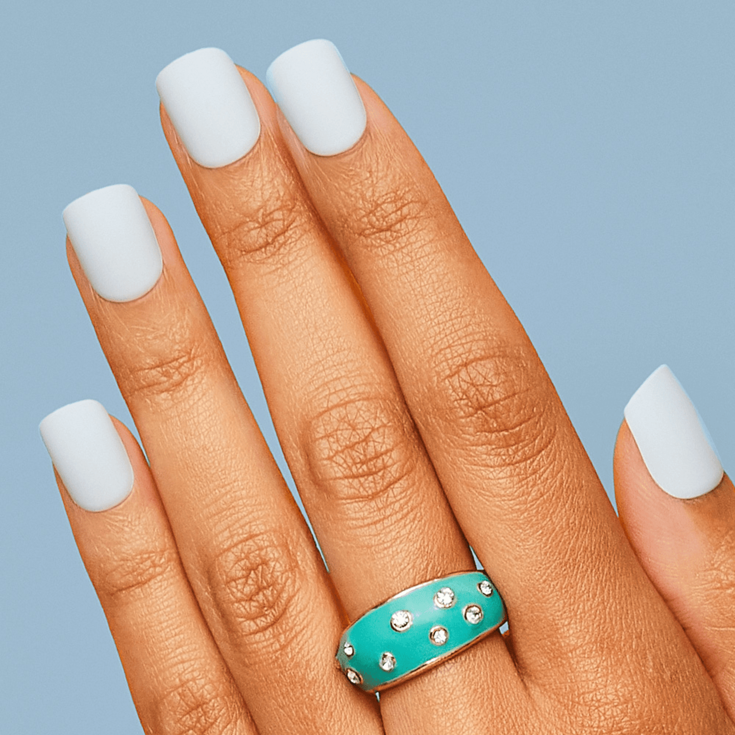 Hand with medium brown skin tone wearing white press-on nails with a matte finish against a light blue background. A turquoise ring with embedded crystals is on the ring finger.