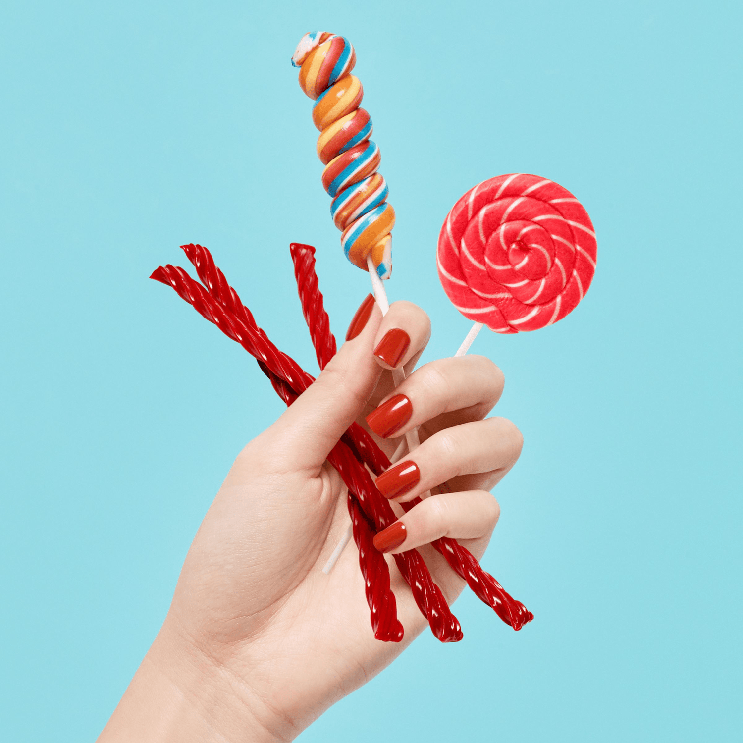 Hand with medium-length square-shaped nails painted in glossy reddish-orange holding colorful candy against a bright blue background.