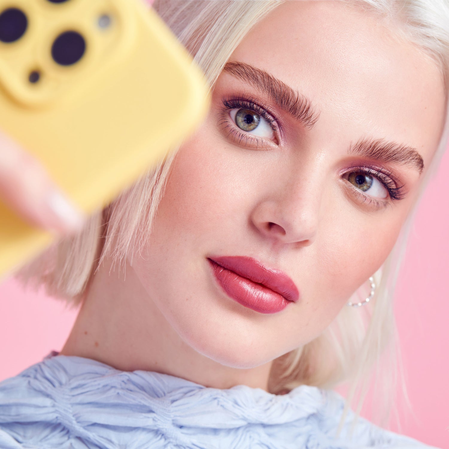 Close-up of a young woman with blonde hair and green eyes wearing natural, voluminous false lashes and mauve eyeshadow, taking a selfie with a yellow phone against a pink background.