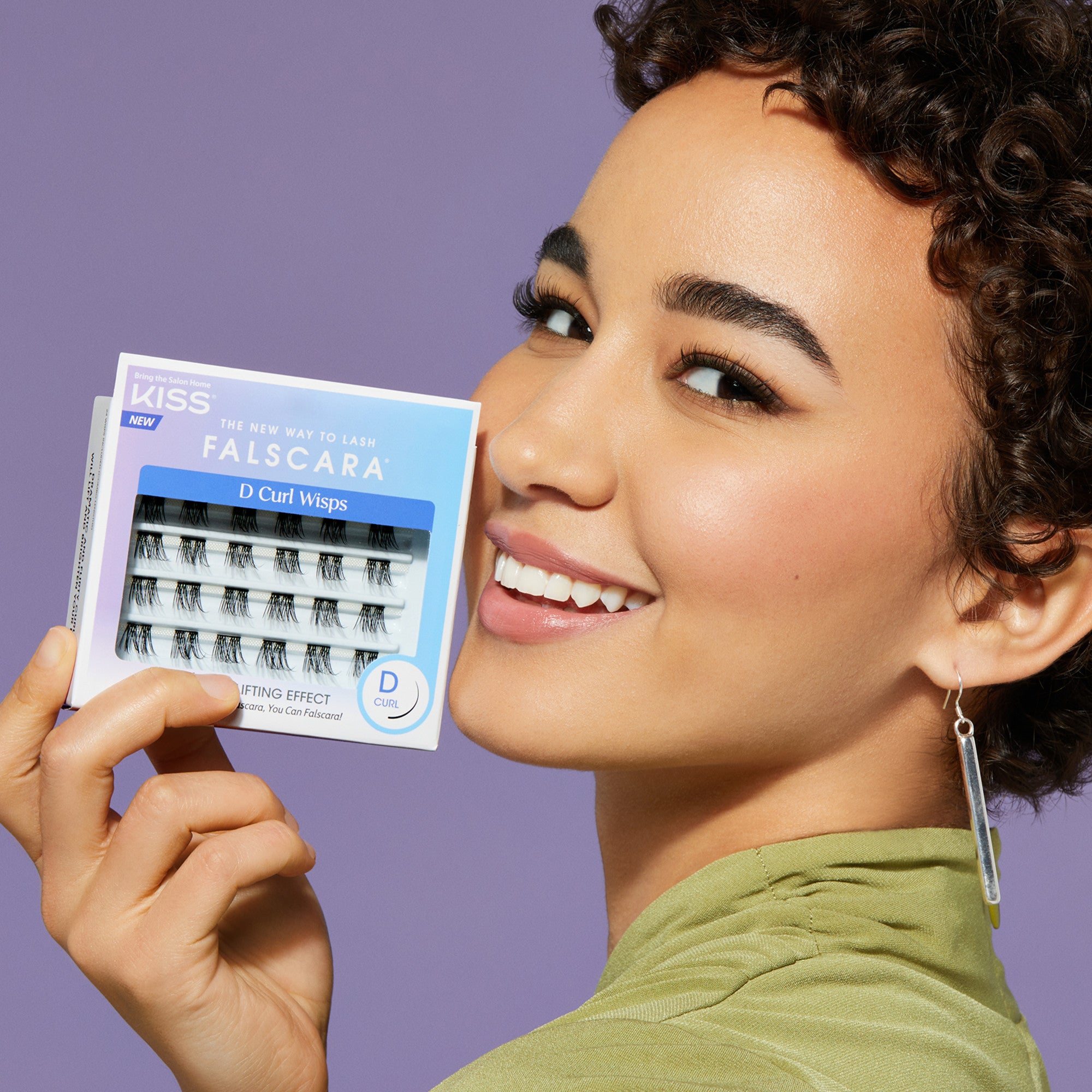Smiling woman with short curly hair and green top holds a box of KISS FALSCARA D Curl Wisps false eyelashes against a purple background.