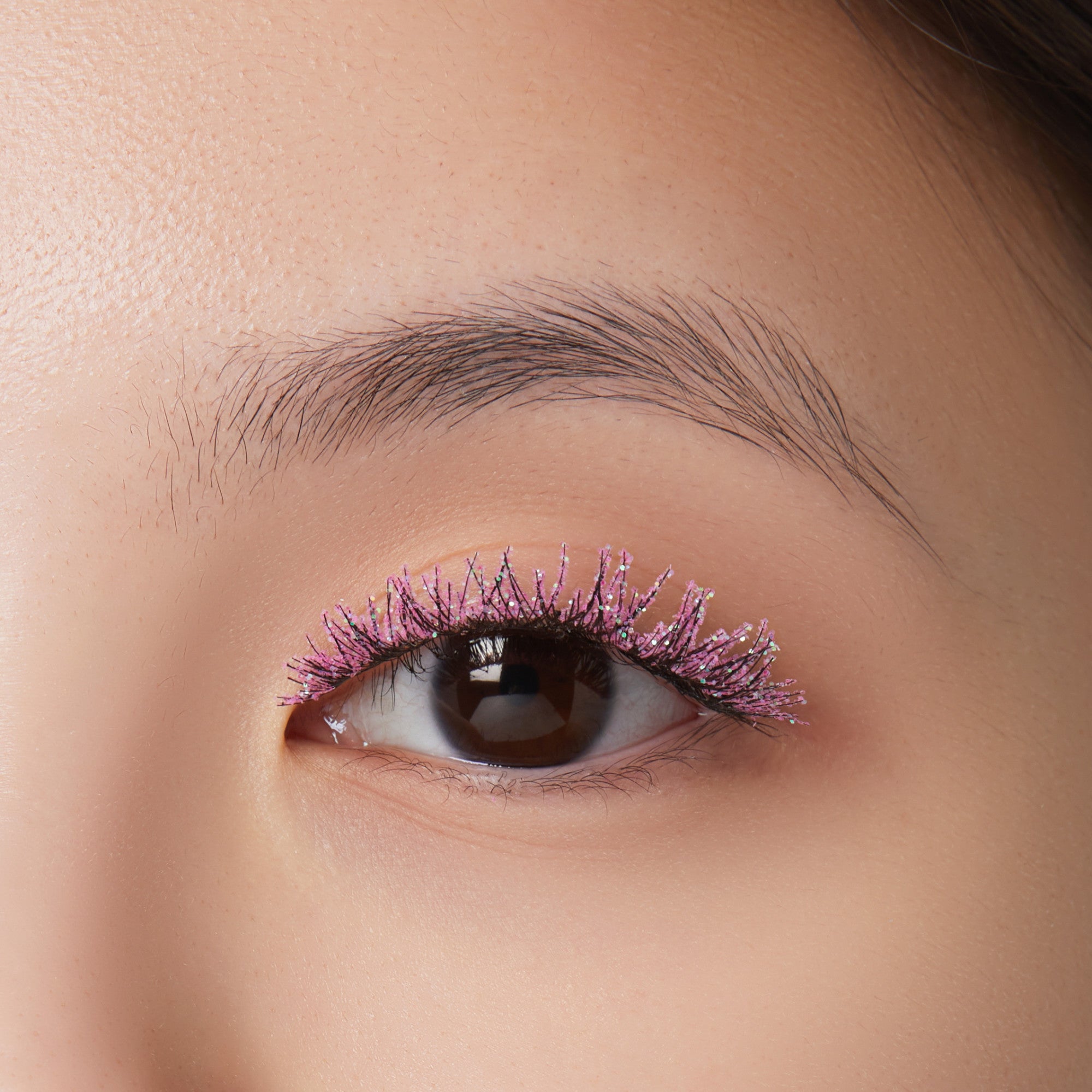 Close-up of a brown eye with long eyelashes coated in sparkling pink glitter mascara, highlighting festive and bold eyelash styling.
