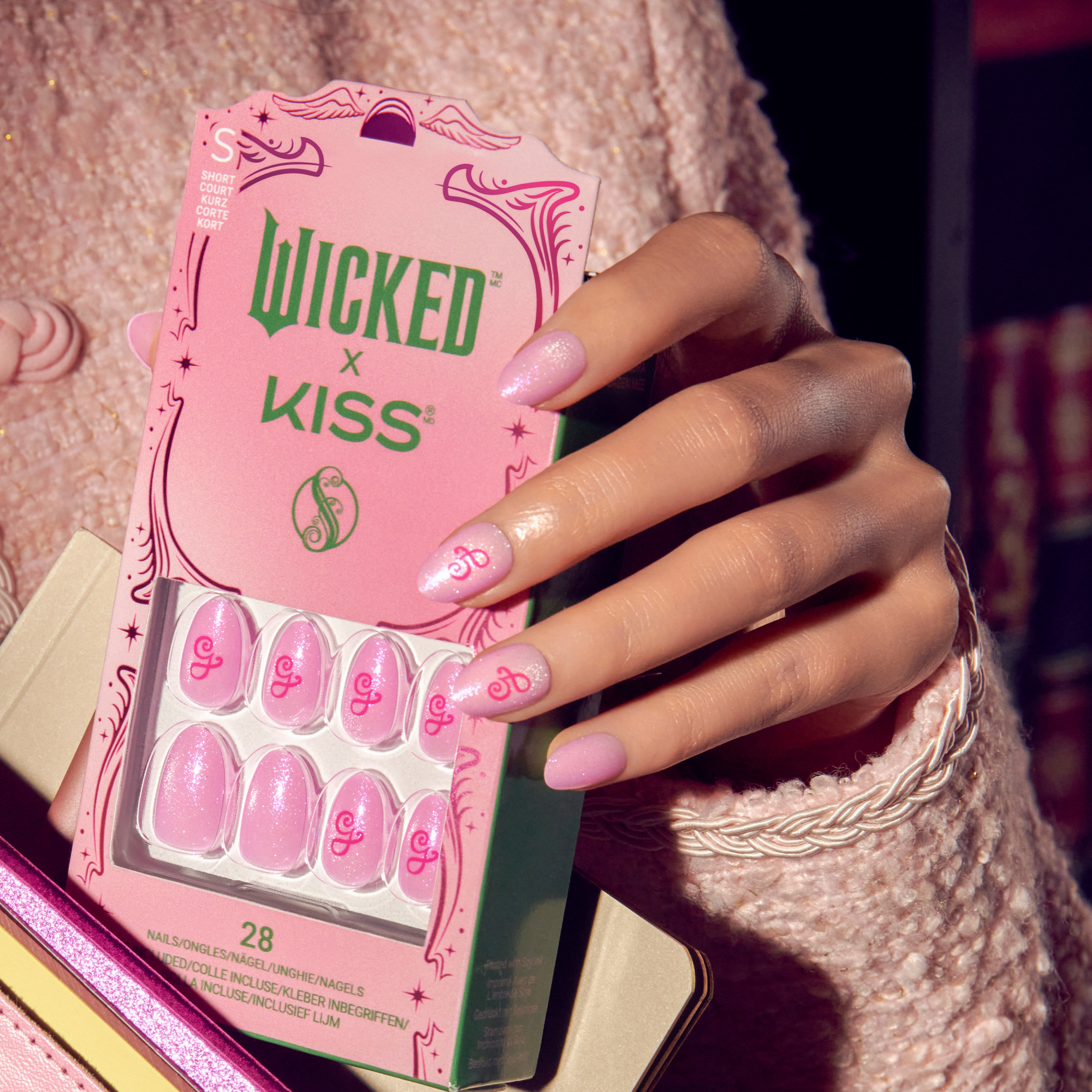 Hand holding a pink package of WICKED X KISS short court press-on nails. The nails are pink with glitter and red stylized \