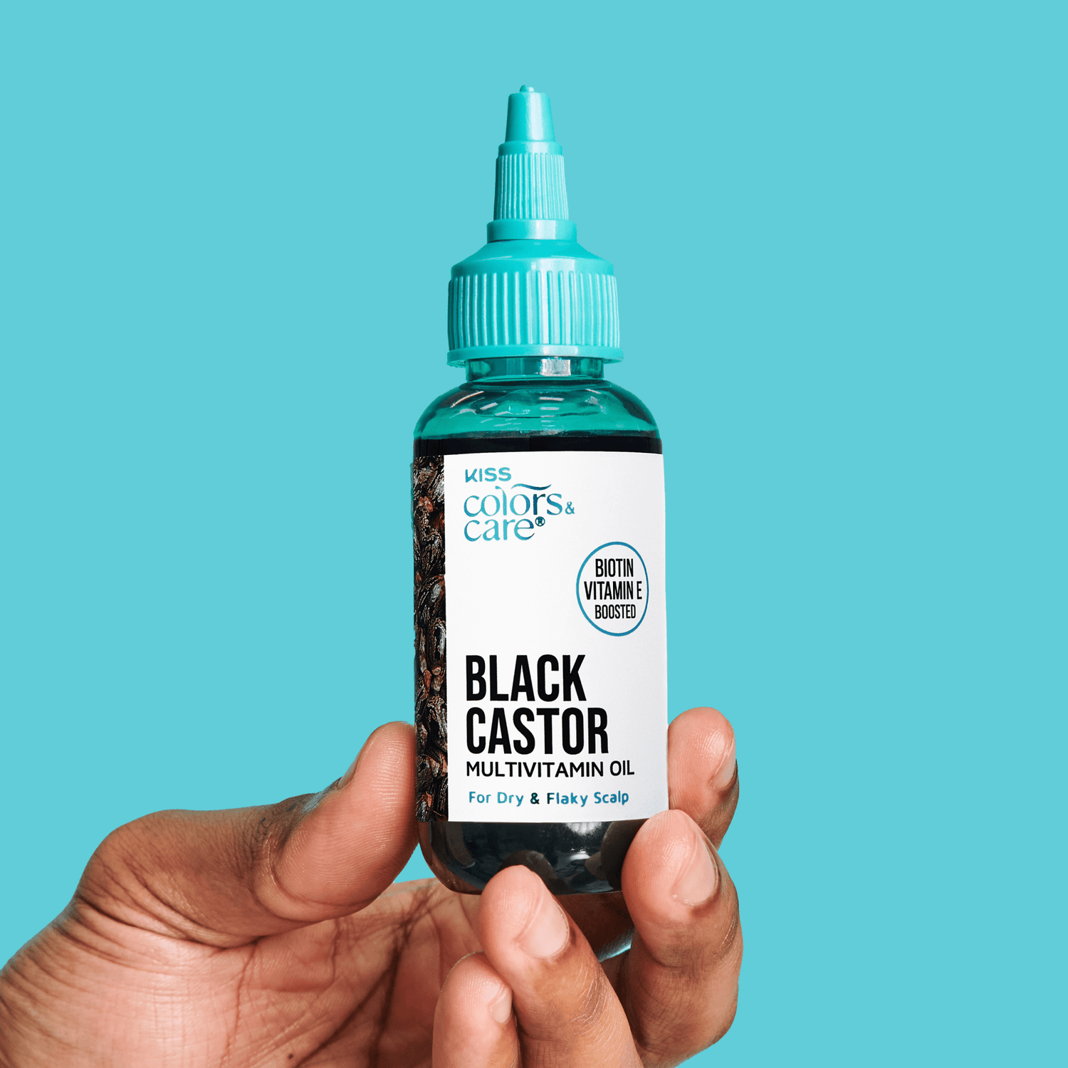 Hand holding a small clear bottle with turquoise dropper cap labeled "KISS Colors &amp; Care Black Castor Multivitamin Oil" for dry and flaky scalp, with biotin and vitamin E boosted.
