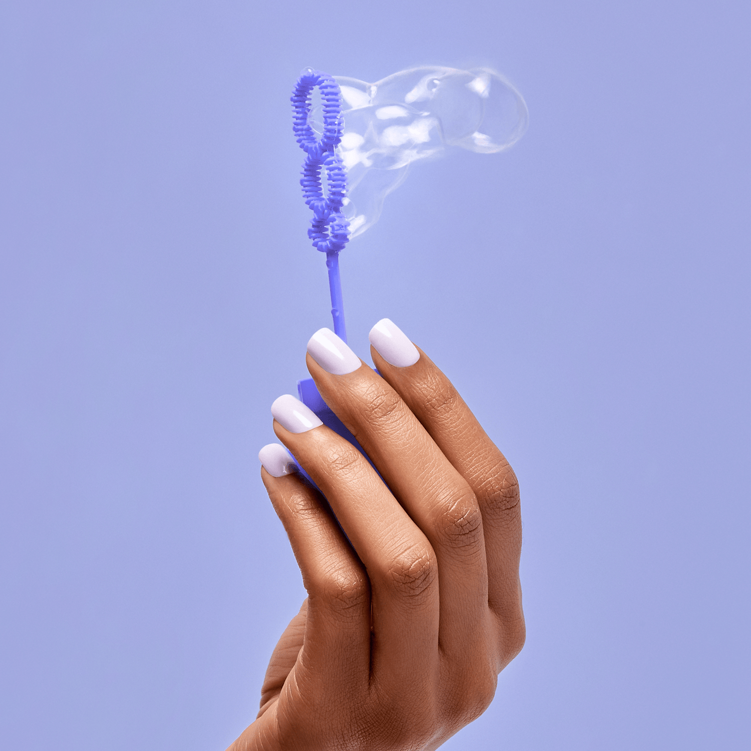 Hand with medium brown skin holding a purple bubble wand, nails painted in a soft pastel purple shade matching the background color.