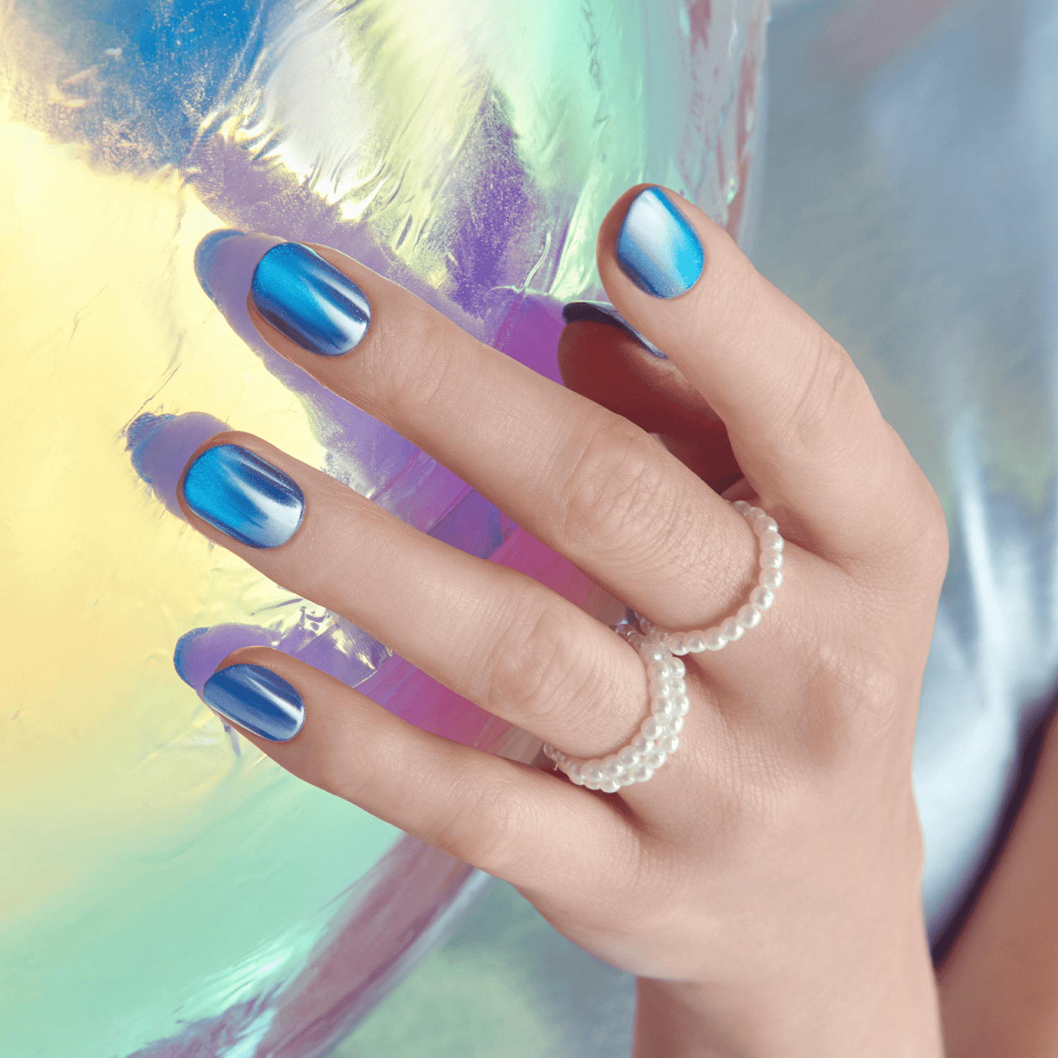 Hand with short press-on nails painted in a metallic blue gradient, wearing two pearl beaded rings, resting against a shiny iridescent surface.