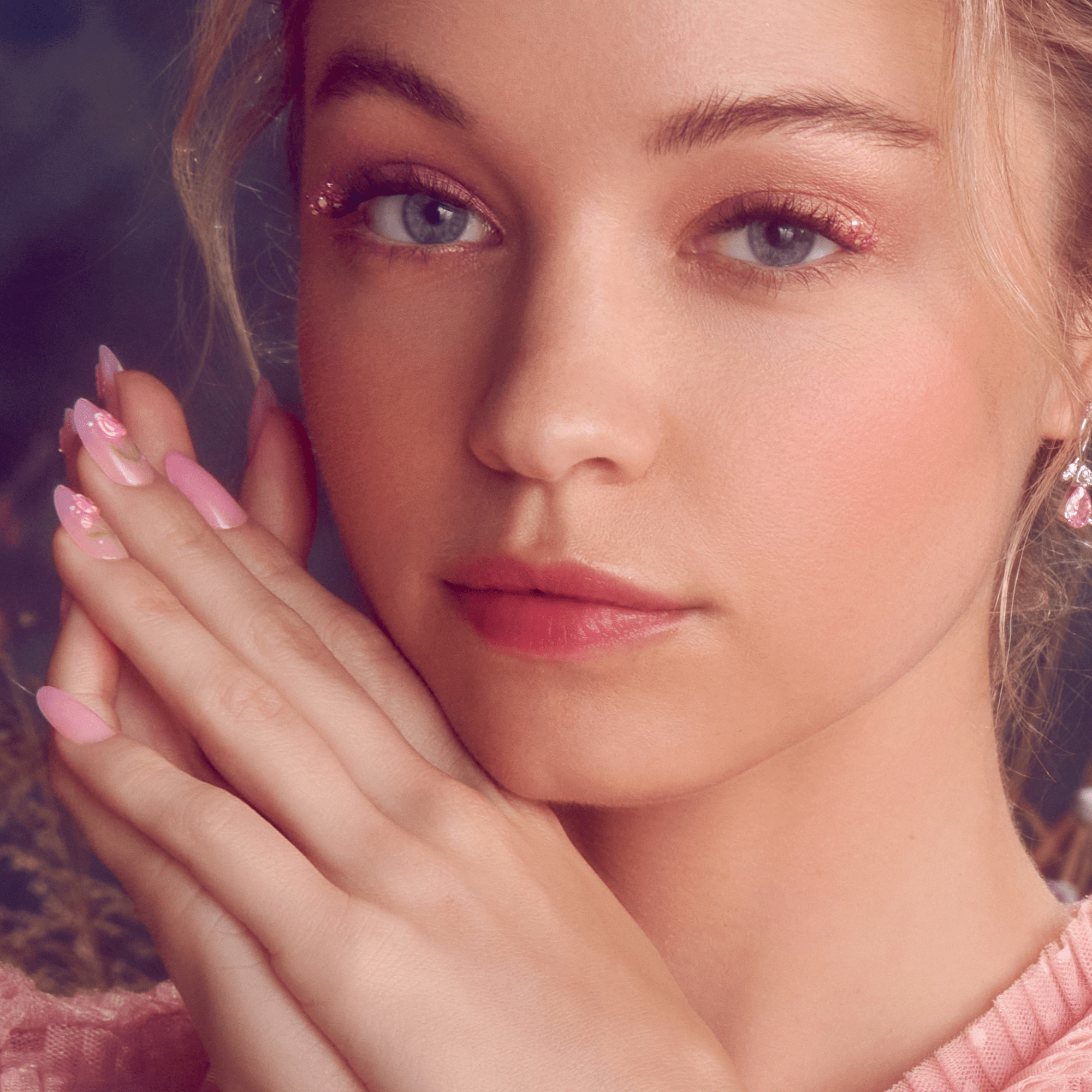 Close-up of a woman with light skin and blue eyes wearing soft pink eyeshadow and lipstick, showing her hands with glossy pink press-on nails featuring delicate floral designs.