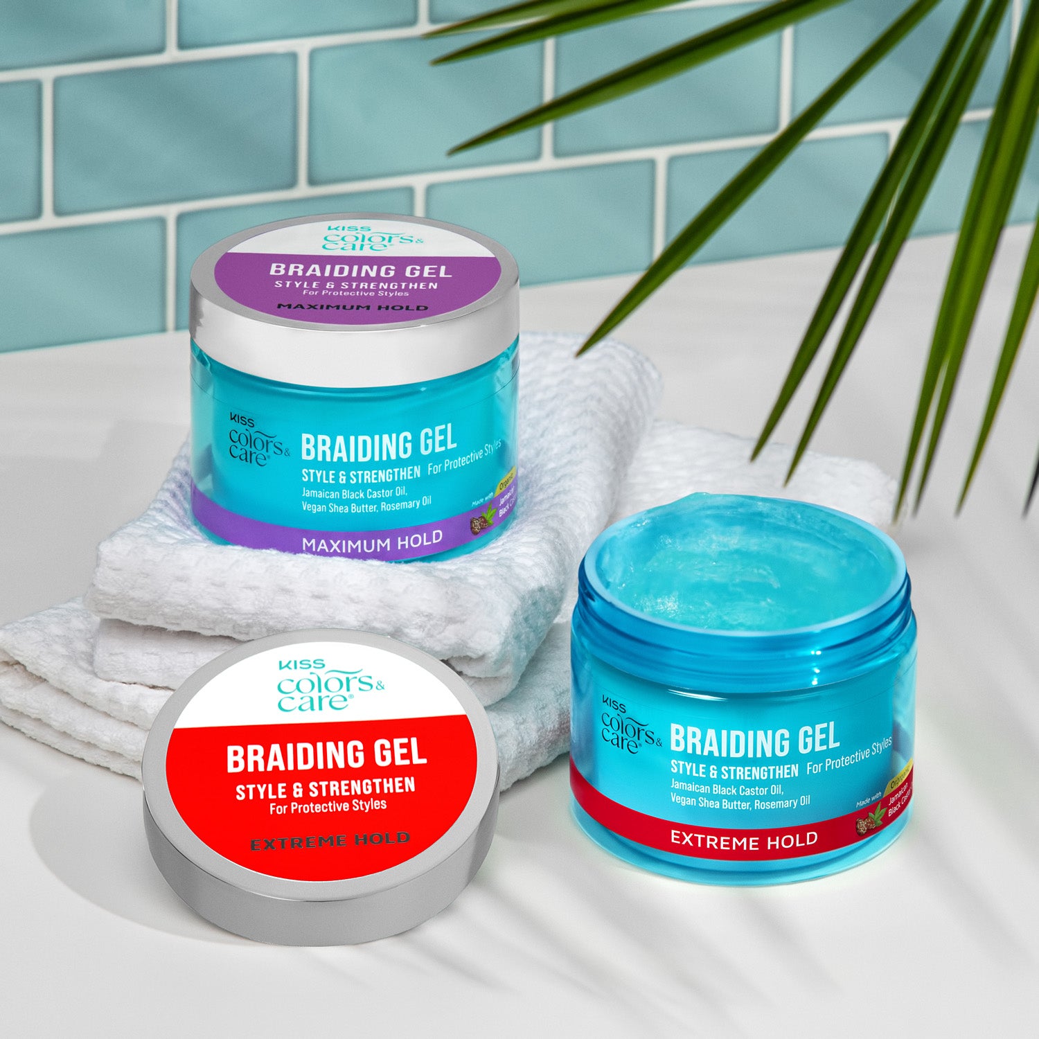 Two blue jars of Kiss Colors &amp; Care Braiding Gel on a white surface, one labeled Maximum Hold with purple accents, the other Extreme Hold with red accents, placed near white towels and a green plant.