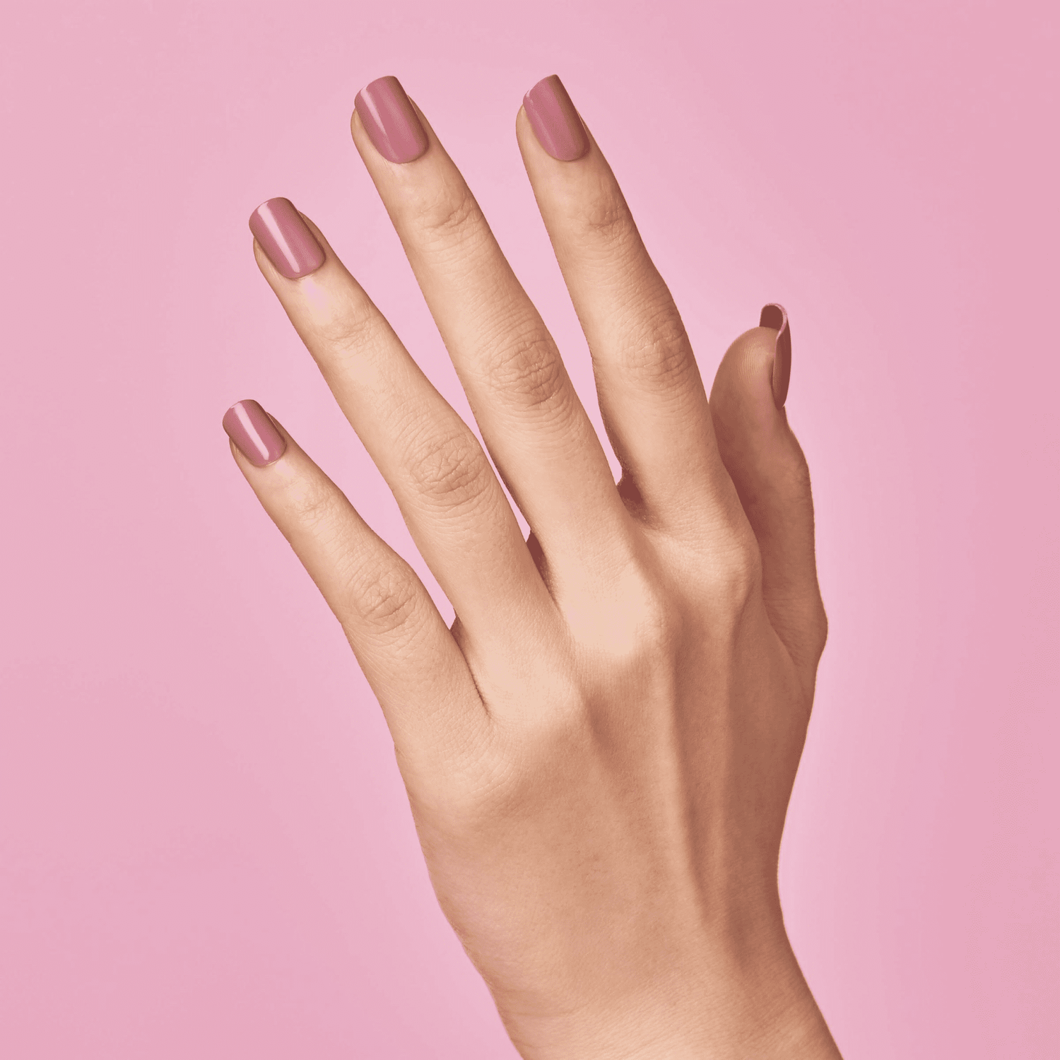 Right hand shown from the back with neatly applied, medium-length press-on nails in a muted petal pink shade against a solid pink background.