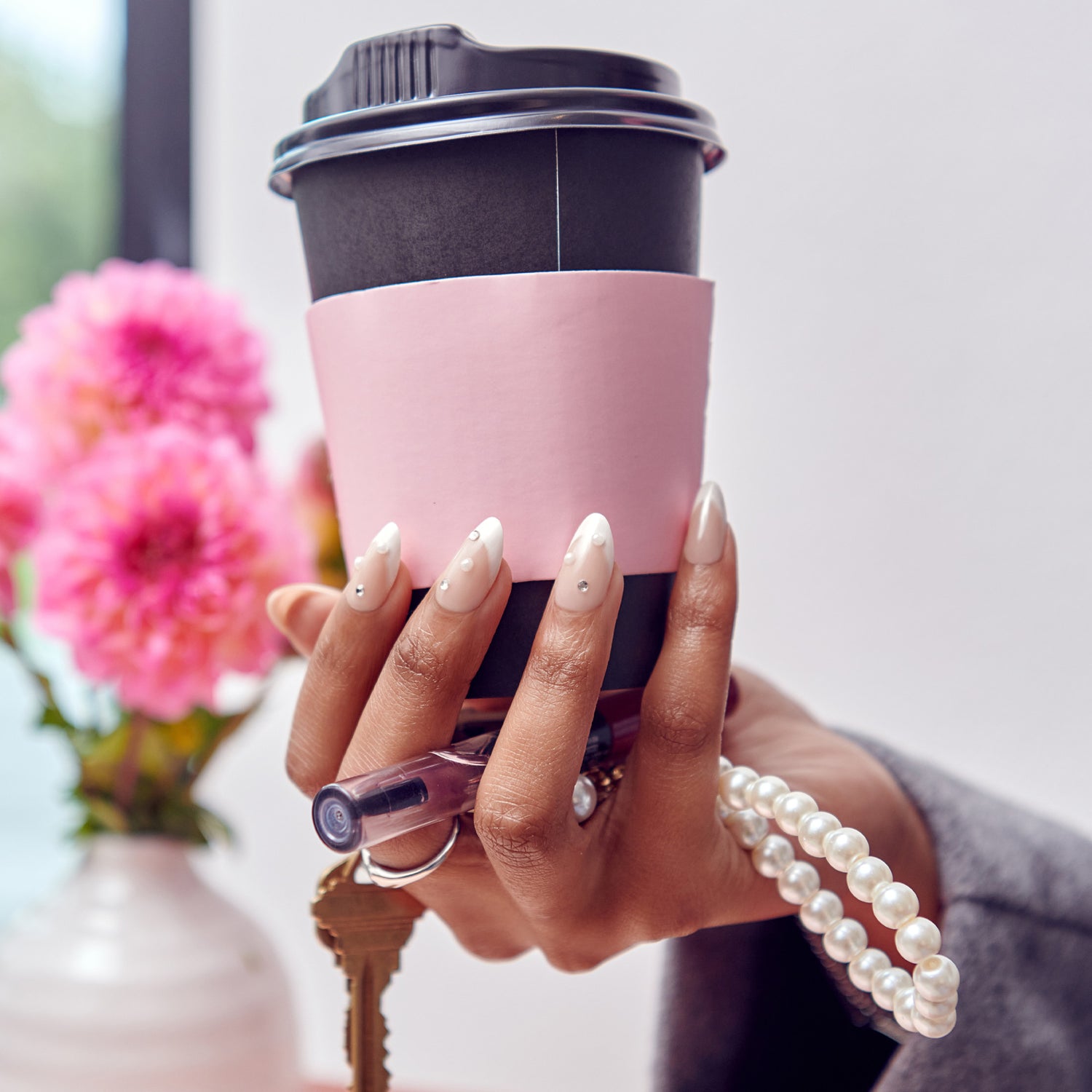 Woman holding pink coffee cup with manicured nails and pearl bracelet near pink flowers