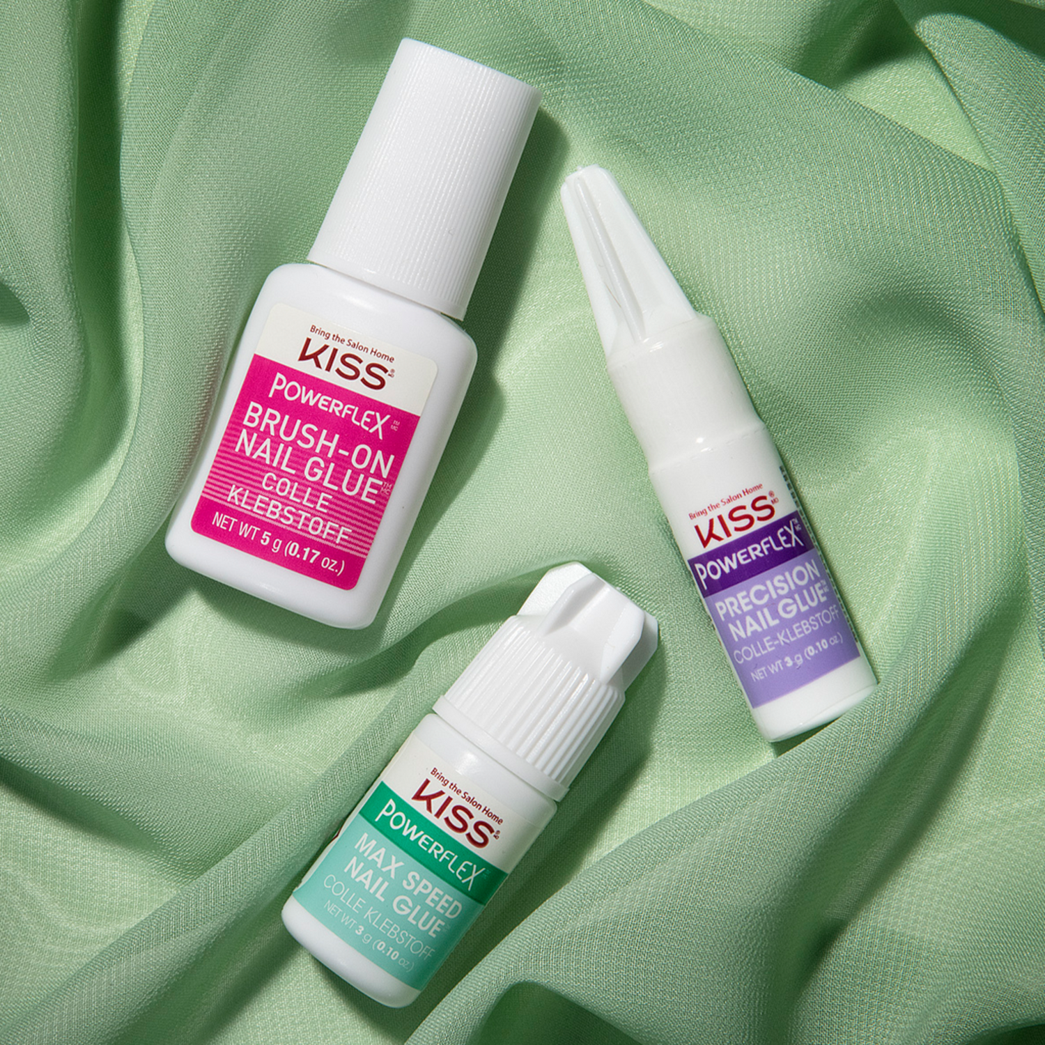 Three small white bottles of KISS PowerFlex nail glue on light green fabric, labeled Brush-On Nail Glue (pink), Max Speed Nail Glue (teal), and Precision Nail Glue (purple).