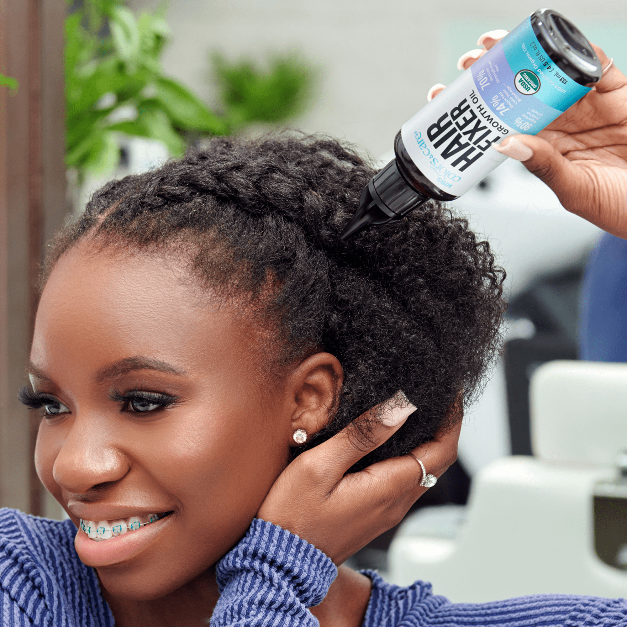 Woman with braided natural hair applying Kiss Colors and Care Hair Fixer Growth Oil from a black-tipped bottle labeled “90% Organic Ingredients” and “Hair Fixer Growth Oil.”