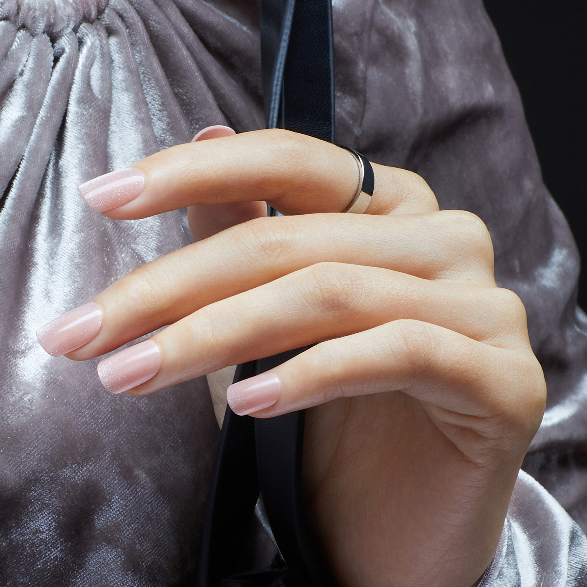 Hand with medium-length, square-shaped press-on nails in a soft pink shade with subtle glitter, worn against a shiny mauve fabric background.