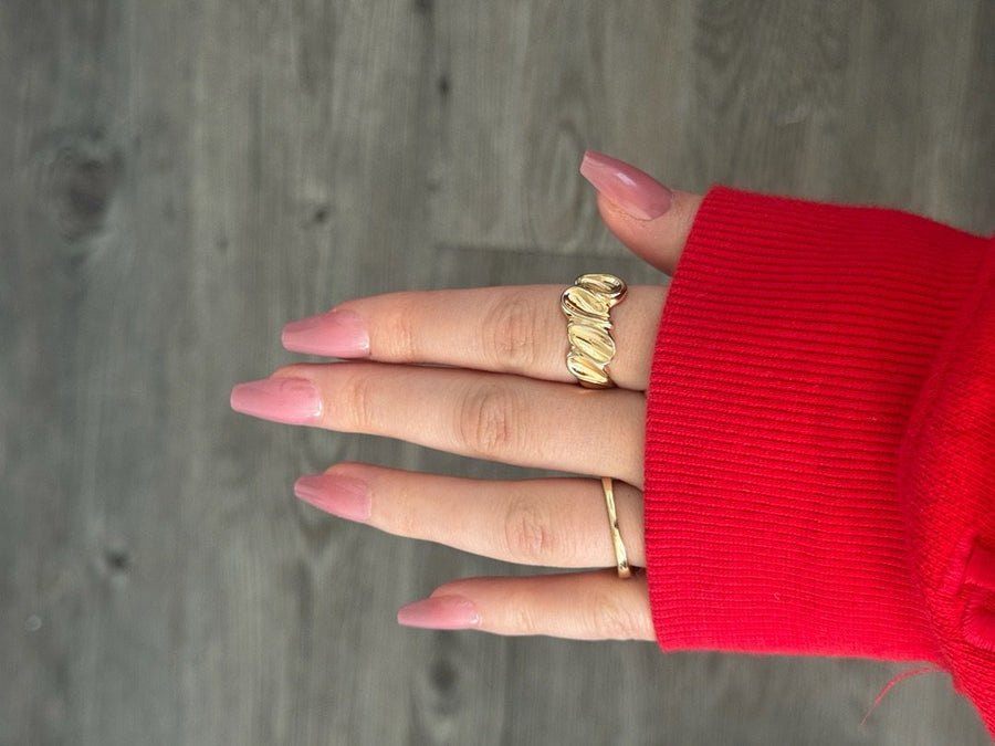 Hand with pink almond nails wearing gold rings and red sweater against gray wood background.