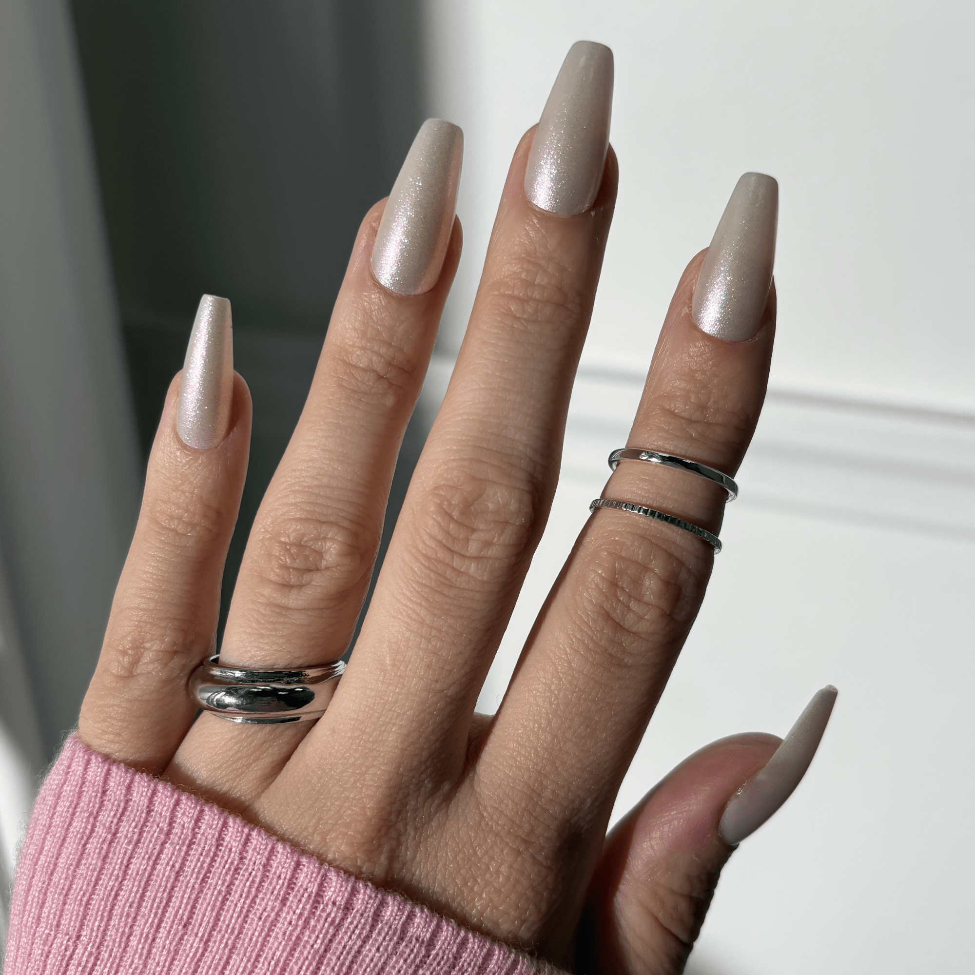 Hand with long, coffin-shaped press-on nails in a pearly light beige shade, worn with silver rings and a pink ribbed sweater sleeve visible.